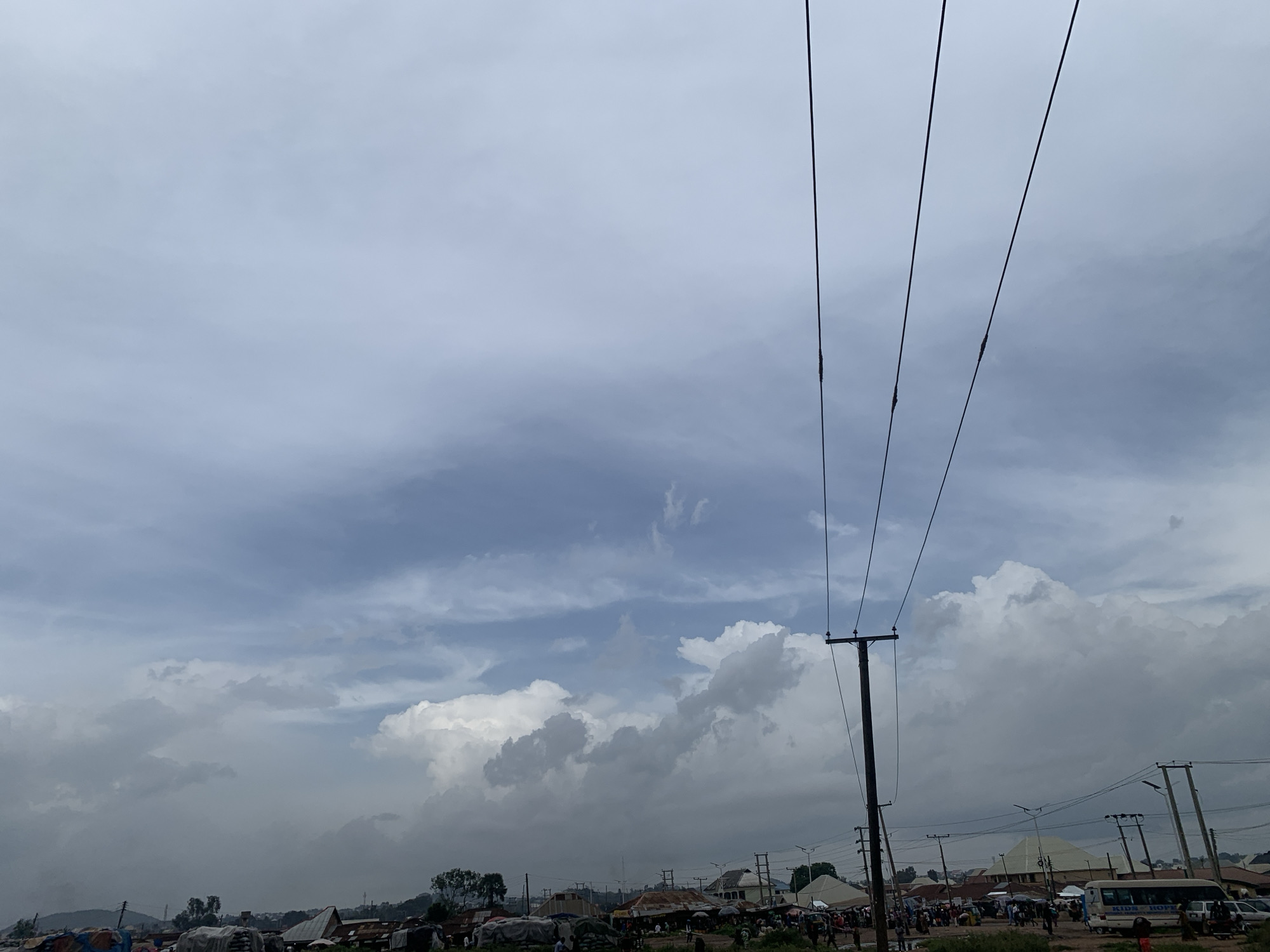 The media file depicts an outdoor scene in Bukuru, Nigeria, under a cloudy sky suggesting it is likely daytime with overcast weather, possibly before or after rain. The foreground is dominated by a utility pole with several power lines stretching diagonally across the frame. In the background, a collection of buildings, some appearing to be market stalls or shops with corrugated iron roofs, are visible. There are also signs of human activity, with numerous people gathered, suggesting a marketplace or a bustling street. The ground appears to be unpaved. A white minibus with "KIDS HOPE" visible on its side is parked among other vehicles, indicating transportation. The overall impression is of a lively, everyday scene in a Nigerian town, characterized by its architecture, infrastructure, and community life.