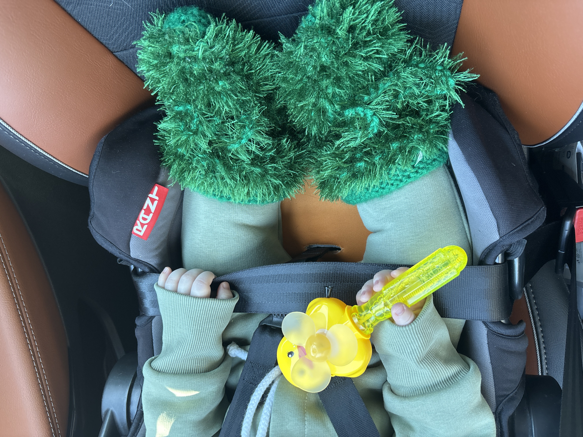 A baby is secured in a car seat, wearing green clothing and fuzzy green booties. The baby is holding a yellow toy with a flower design. The car seat is positioned inside a vehicle with brown leather interior. The city of Chaikovsky, Russia is not visually discernable in this image.