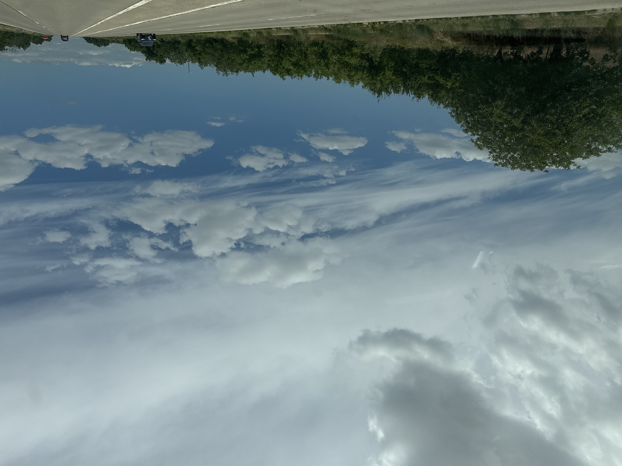 This is a photograph of a highway under a cloudy sky in Horesti, Moldova. The image is taken from a moving vehicle and captures a landscape view. The foreground shows a paved road with white lane markings, indicating a multi-lane highway. On the left side of the road, there is a dense line of green trees and vegetation. Further back, the terrain appears to be slightly elevated with more trees.
The sky is a prominent feature, filled with a variety of clouds. Some clouds are dense and grey, suggesting potential for rain, while others are lighter and scattered, allowing patches of blue sky to show through. Wispy cirrus clouds are also visible, streaking across the sky. The lighting suggests it is daytime, possibly late morning or early afternoon, with diffused sunlight.
In the distance, two vehicles are visible on the highway. One is a dark-colored SUV in the left lane, and another vehicle, possibly a sedan, is further ahead in the right lane. The overall scene conveys a sense of travel or transit through a rural or semi-rural environment. There are no discernible people visible in the image, and no specific landmarks of Horesti are identifiable from this perspective.
