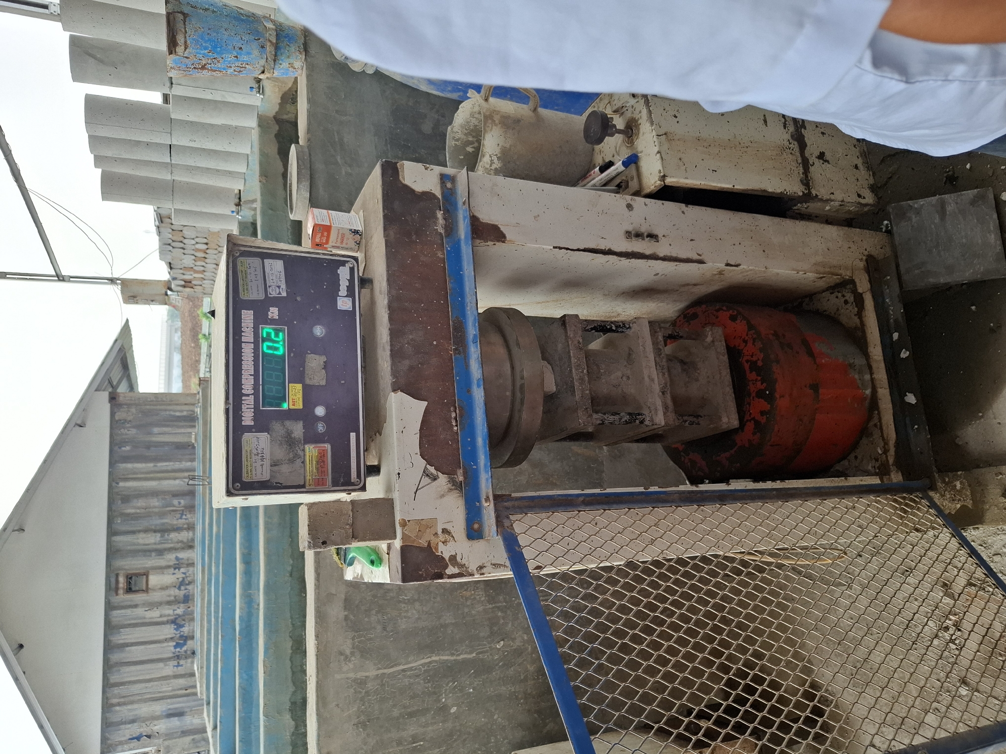 In Kutasirna, Indonesia, a concrete compression testing machine stands in a dimly lit outdoor setting. The machine, a bulky, weathered metal device with a digital display showing "0.2", is positioned in front of rows of concrete cylinders. A wire mesh gate, slightly ajar, protects the lower portion of the machine. To the right, a person in a white shirt is partially visible, suggesting an industrial or laboratory environment. The background features a simple building with corrugated walls and a sloped roof. The overall scene conveys a sense of ongoing construction or material testing.
