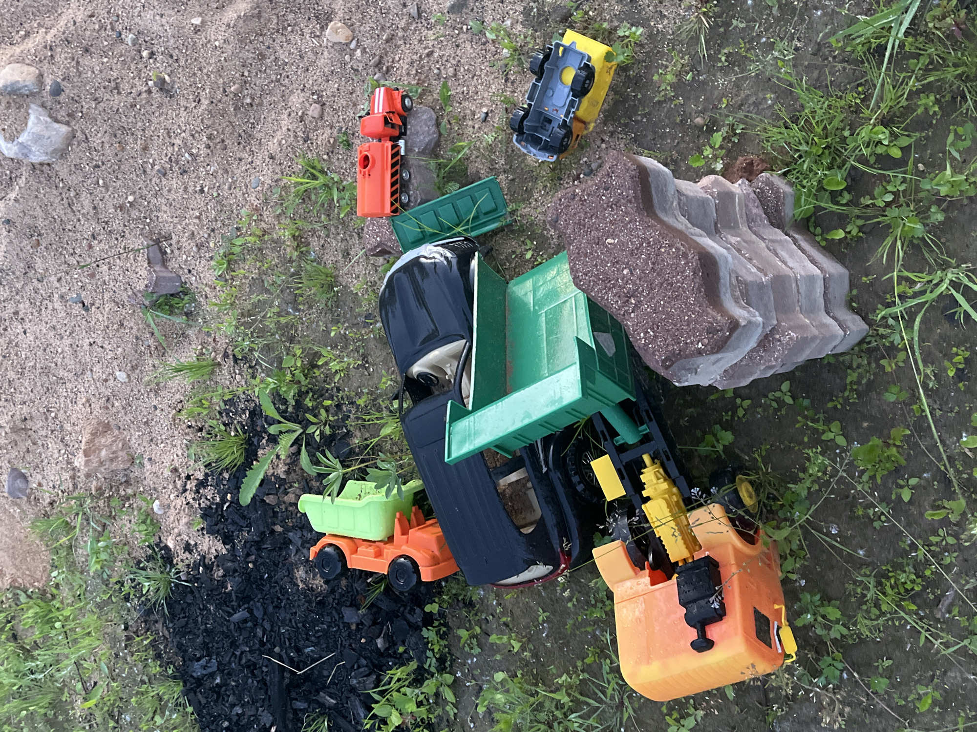An outdoor, high-angle, eye-level shot shows several overturned toy trucks scattered on a patch of dirt, grass, and sand. In the upper left corner, a pile of black, burnt debris sits amongst green grass and weeds. To the right of the debris, an orange and green toy truck is lying on its side. A black toy SUV with a green dump bed is in the center of the frame. To the right of the SUV, an orange toy tanker truck sits on top of a grey rock. Below the tanker truck, a yellow toy truck with a grey chassis is overturned. In the lower right of the frame, a stack of interlocking paving stones sits in the dirt. The overall impression is of a disused, neglected area.