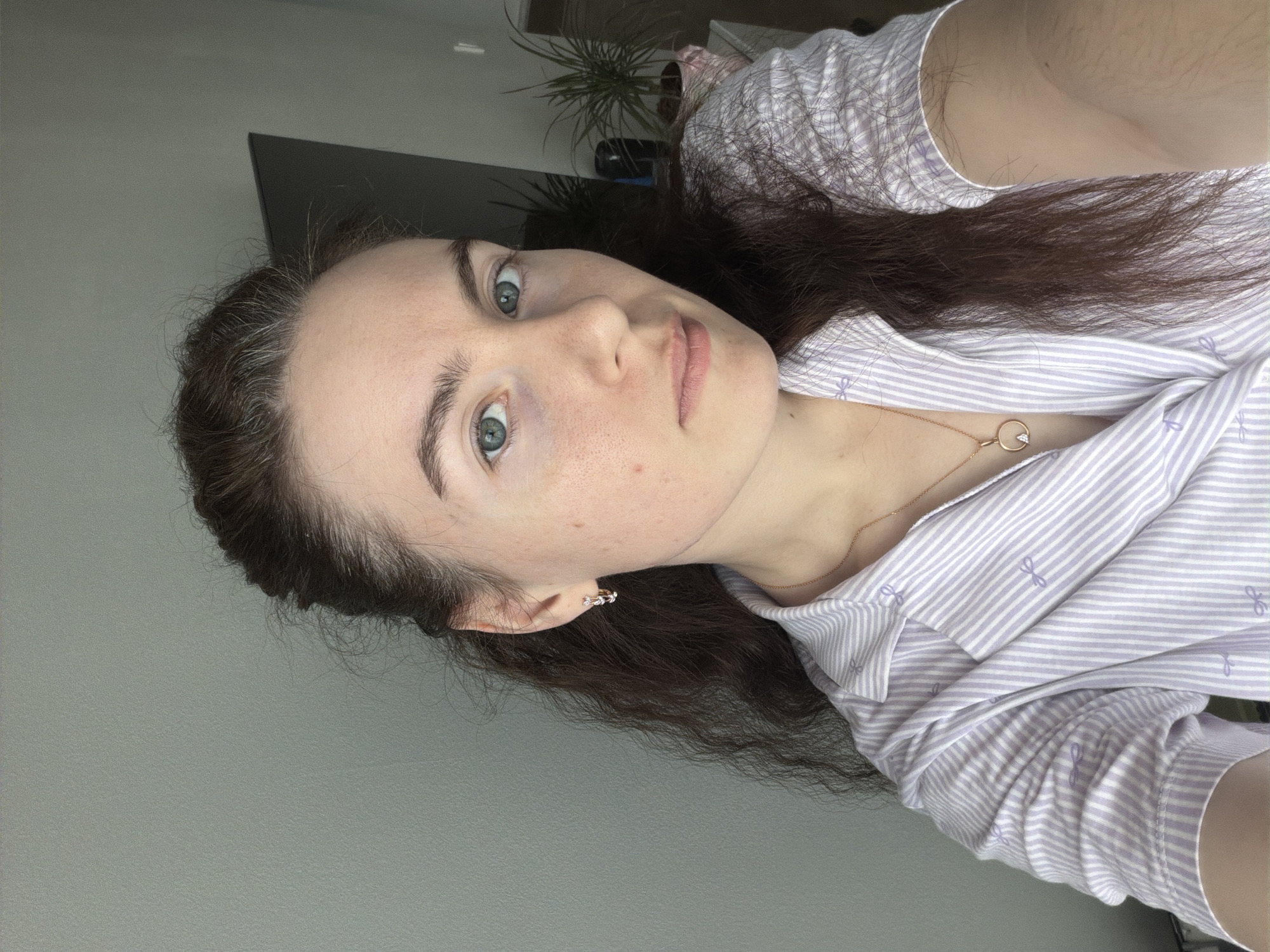 A selfie of a Caucasian woman with long brown hair, wearing a light purple and white striped shirt and a gold necklace with a circular pendant. She has light skin and blue eyes. An earring is visible in her left ear. The background is a grey wall and partially visible TV screen. A potted plant is slightly visible behind her. There is no verifiable evidence to support that this image was taken in Yanino-1, Russia.