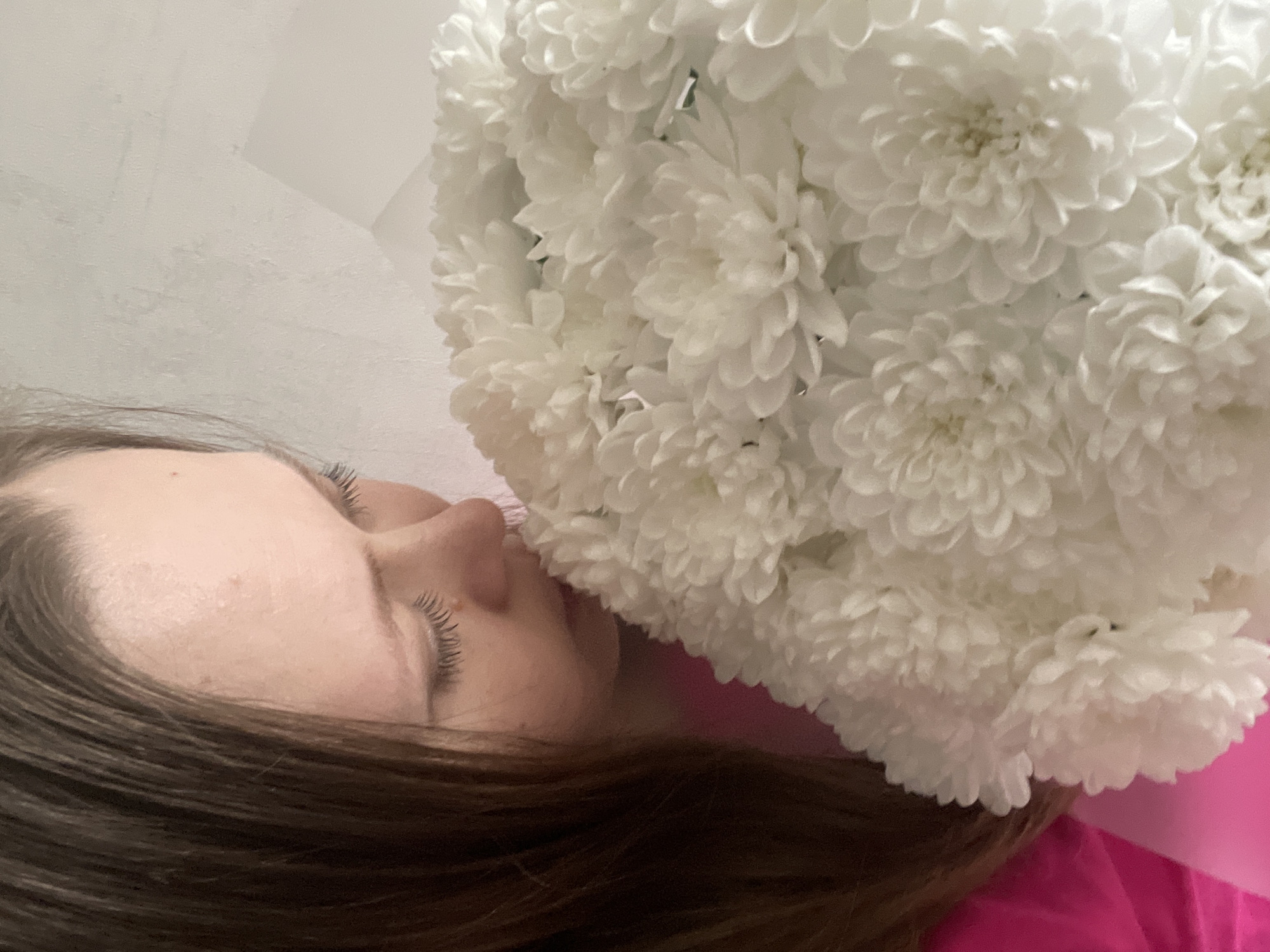 A woman with brown hair is lying down, her eyes closed, with a large bouquet of white chrysanthemum flowers positioned near her face. She is wearing a pink garment. The background appears to be a white wall. It's impossible to independently verify the location in the city of Chaikovsky, Russia.