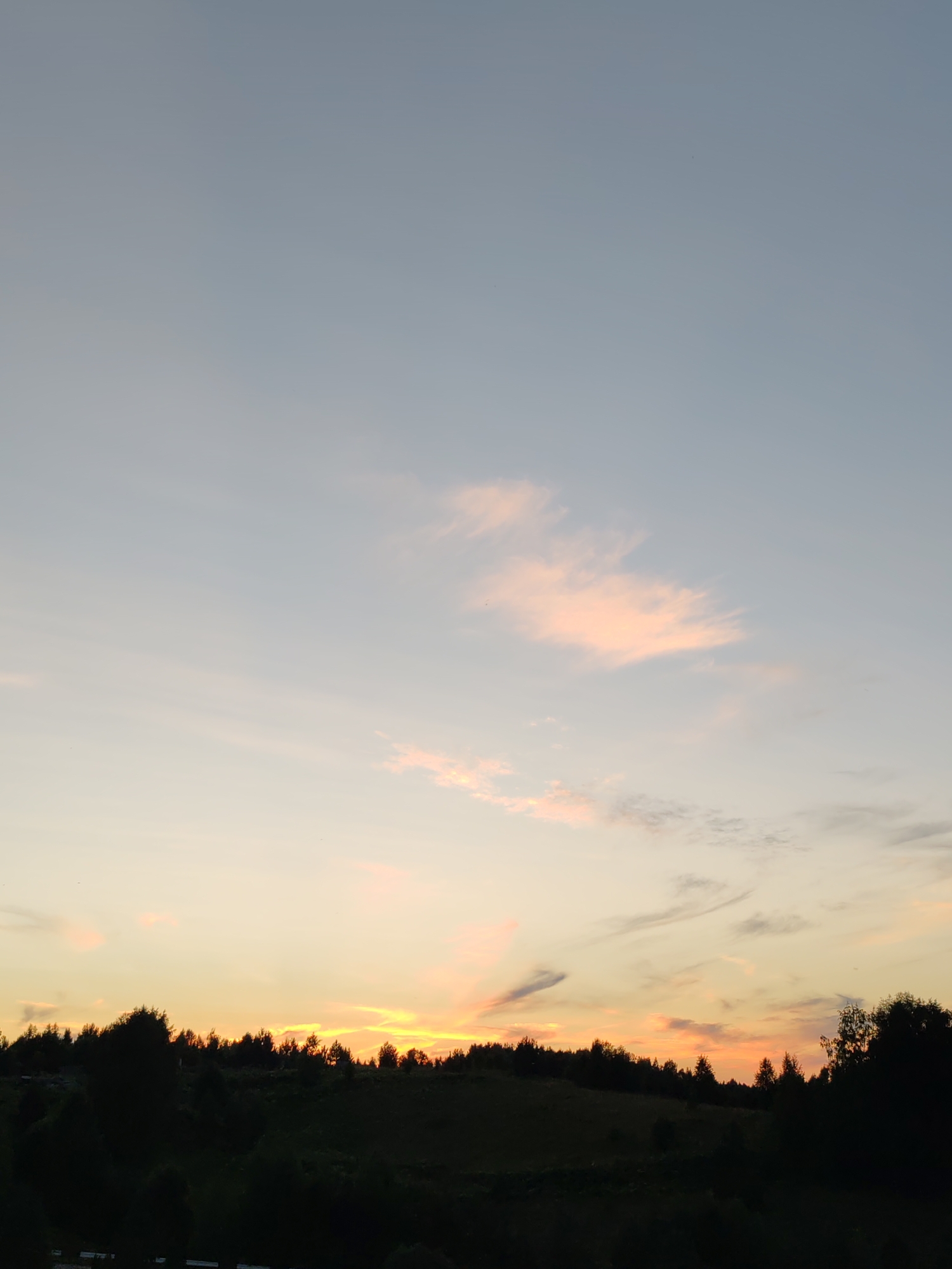 This is a wide shot of a sunset over a rural landscape. The sky is a pale blue fading to yellow and orange near the horizon. The sun is setting behind a silhouetted treeline that stretches across the middle ground. The treeline is composed of various evergreen and deciduous trees. In the foreground, there is a dark silhouette of a hill with some faint details of vegetation. The overall impression is one of a peaceful, natural setting at dusk. The image does not contain any people or identifiable structures. The location is described as Чухлэм, Russia, implying a rural, possibly forested, area.