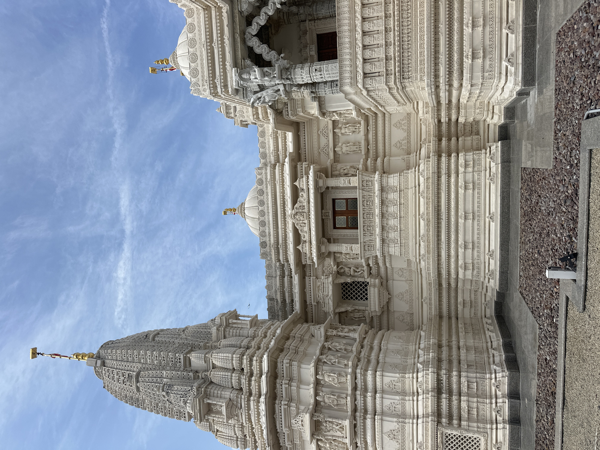 The image shows the BAPS Shri Swaminarayan Mandir, a Hindu temple located in Bartlett, Illinois, USA. The temple is constructed from white marble and features intricate carvings and sculptures. The foreground of the image shows a gravel pathway leading to the temple. The temple itself is the main subject, with its ornate towers and domes. The sky is blue with scattered clouds. The overall scene is serene and spiritual.
