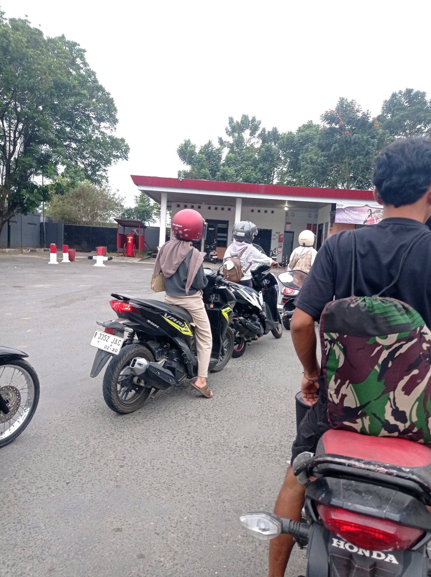 In the city of Bojong, Indonesia, on an overcast day, a line of motorcycles is waiting at a gas station. The foreground features the back of a person wearing a black t-shirt and dark shorts, with a camouflage backpack slung over their shoulder. Their right leg is visible, resting on the seat of a red and black Honda motorcycle. To the left of this motorcycle, the front wheel of another motorcycle is visible.
Further into the scene, a person in a red helmet is seated on a black motorcycle, facing away from the camera. They are wearing a grey hoodie and light-colored pants, with a beige bag over their shoulder. To their right, another person on a motorcycle is partially visible, wearing a white helmet and a light-colored top. In the background, the gas station structure with a red roof and white pillars is present. Several red and white bollards and a red fire extinguisher cabinet are also visible in the outdoor area of the gas station. Trees with green foliage form the backdrop. The road surface is asphalt with visible texture.