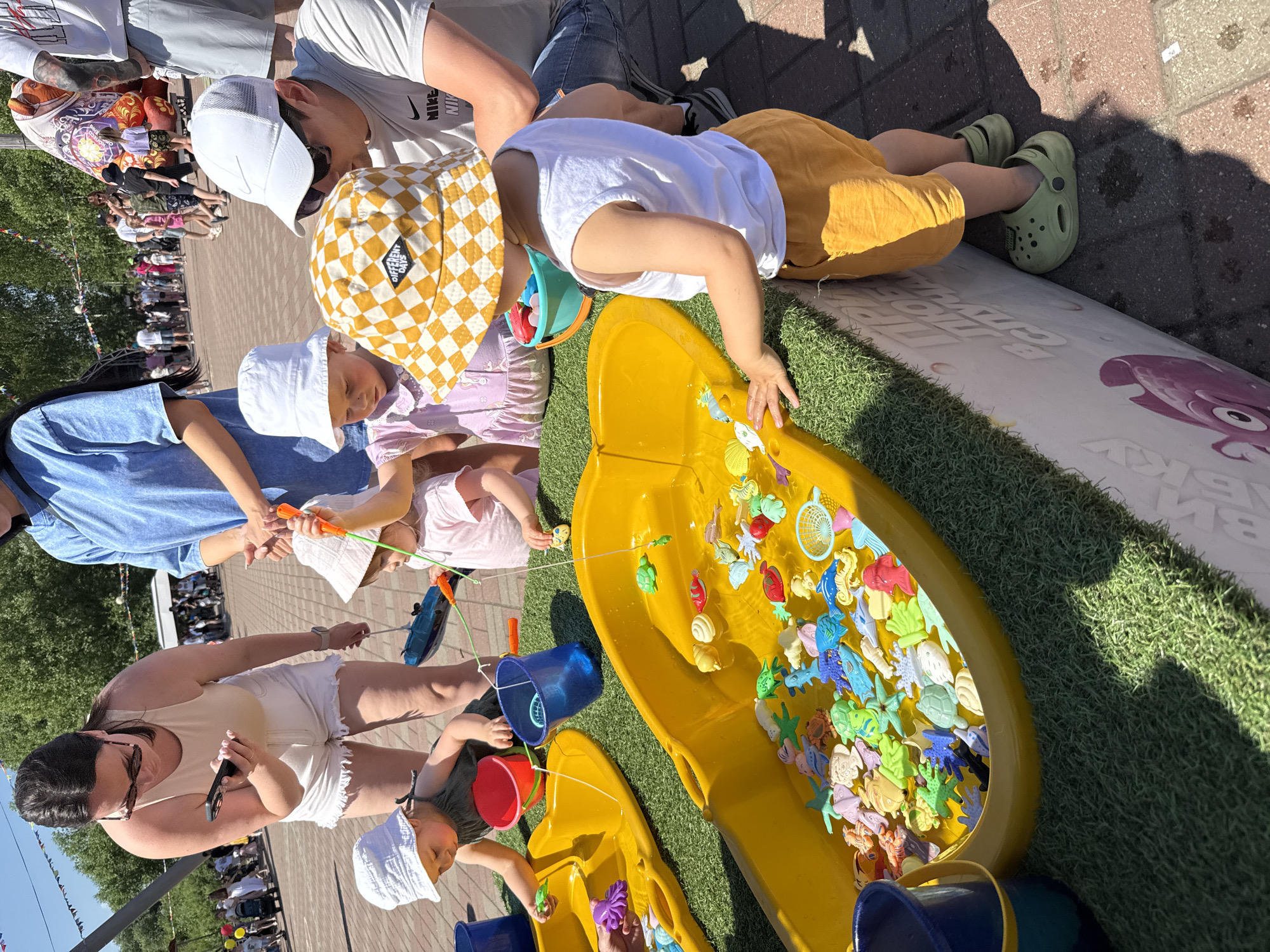 At an outdoor public event in Zavolzhye, Russia, a group of young children are engaged in a fishing game. The game features a large yellow plastic trough filled with water and numerous small, colorful plastic sea creatures, including fish, turtles, and starfish. Each child is equipped with a fishing rod, which appears to be a stick with a string and a magnet at the end, used to attract the magnetic toys.
A woman in a beige tank top and white shorts supervises the children, holding a smartphone in her hand. Another adult, wearing a blue shirt, stands behind her. A man in a white t-shirt, white cap, and sunglasses is crouched down, assisting one of the children. The children are dressed casually for warm weather, with two wearing bucket hats. One child wears a green tank top and yellow shorts, while another wears a pink dress.
The setup is on a patch of artificial green turf. In the background, a crowd of people, including adults and children, are milling about. Various decorations, possibly flags or banners, are visible overhead. The overall setting suggests a festive atmosphere at a community gathering or fairground. The ground is paved with brick-like stones.