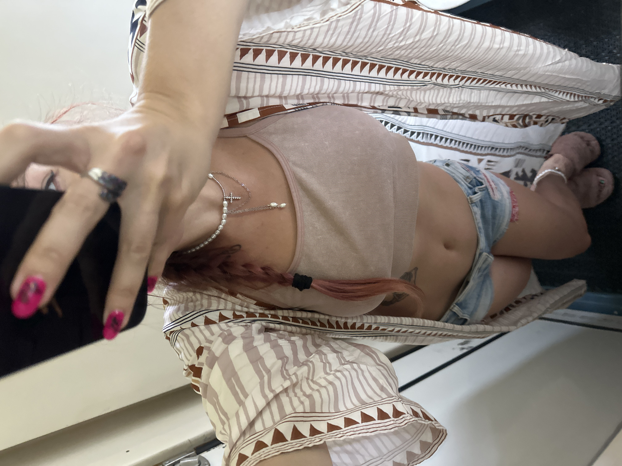 A person, with pink hair braided to their side, is taking a selfie in what appears to be a bathroom. They are wearing a tan crop top, denim shorts, and a patterned kimono-style robe. The person has bright pink painted nails and is wearing jewelry, including a pearl necklace with a cross pendant and rings. They are also wearing fuzzy slippers. The background is mostly a light-colored wall and a dark floor. The image is taken from a high angle. There is no visible indication of the city of Покровского Торфоучастка поселок, Russia.