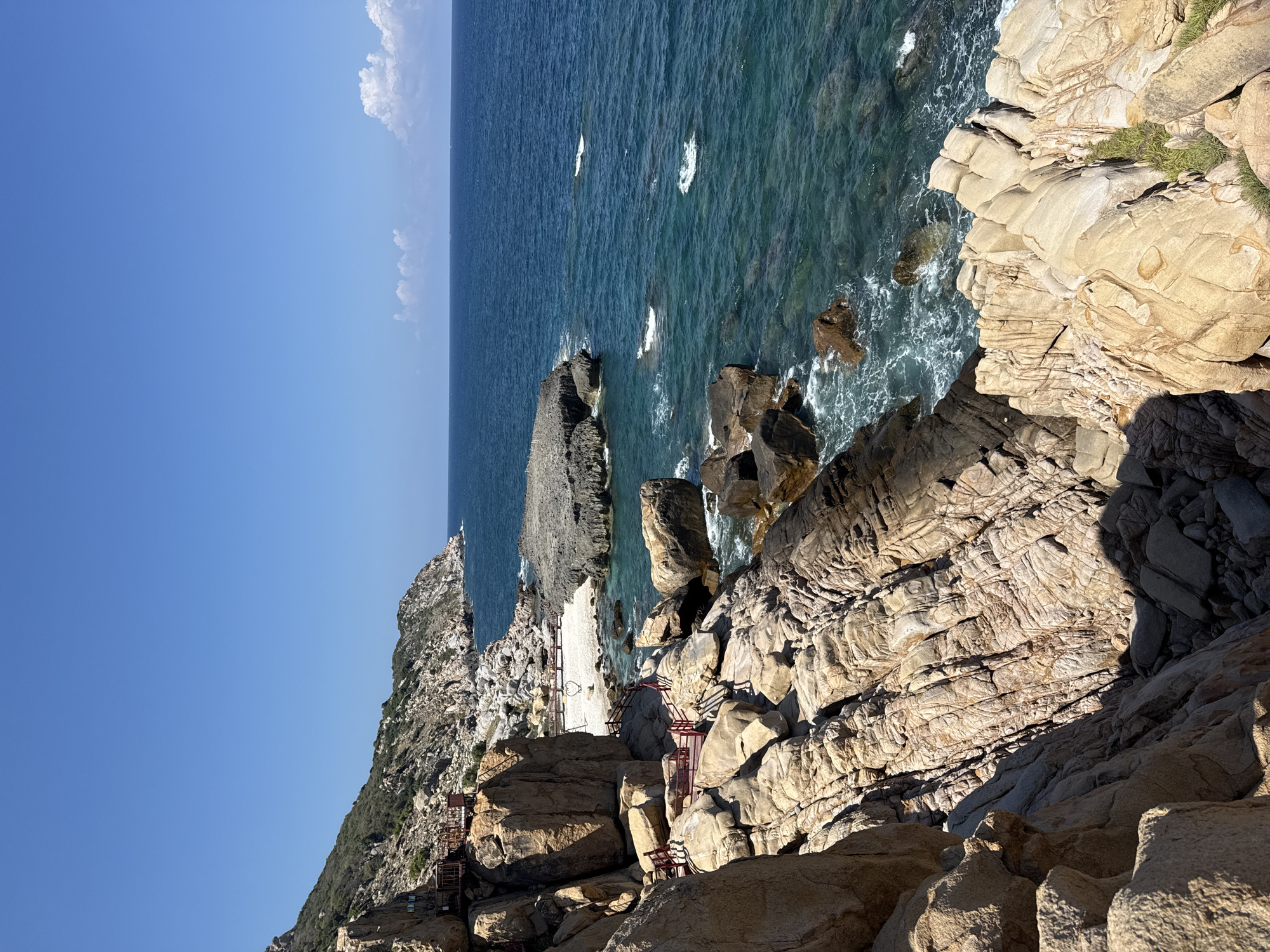A scenic view of the rocky coastline in Vinh Hai, Vietnam, taken on a sunny day with clear blue skies. The turquoise sea crashes against the rugged cliffs made of light brown and grey rocks. A series of red metal staircases are built into the rocks to provide access to a small beach visible in the middle ground. Vegetation is sparse, with small patches of green growth on the rocks. The photograph's perspective is from above, looking down at the sea and the coast.