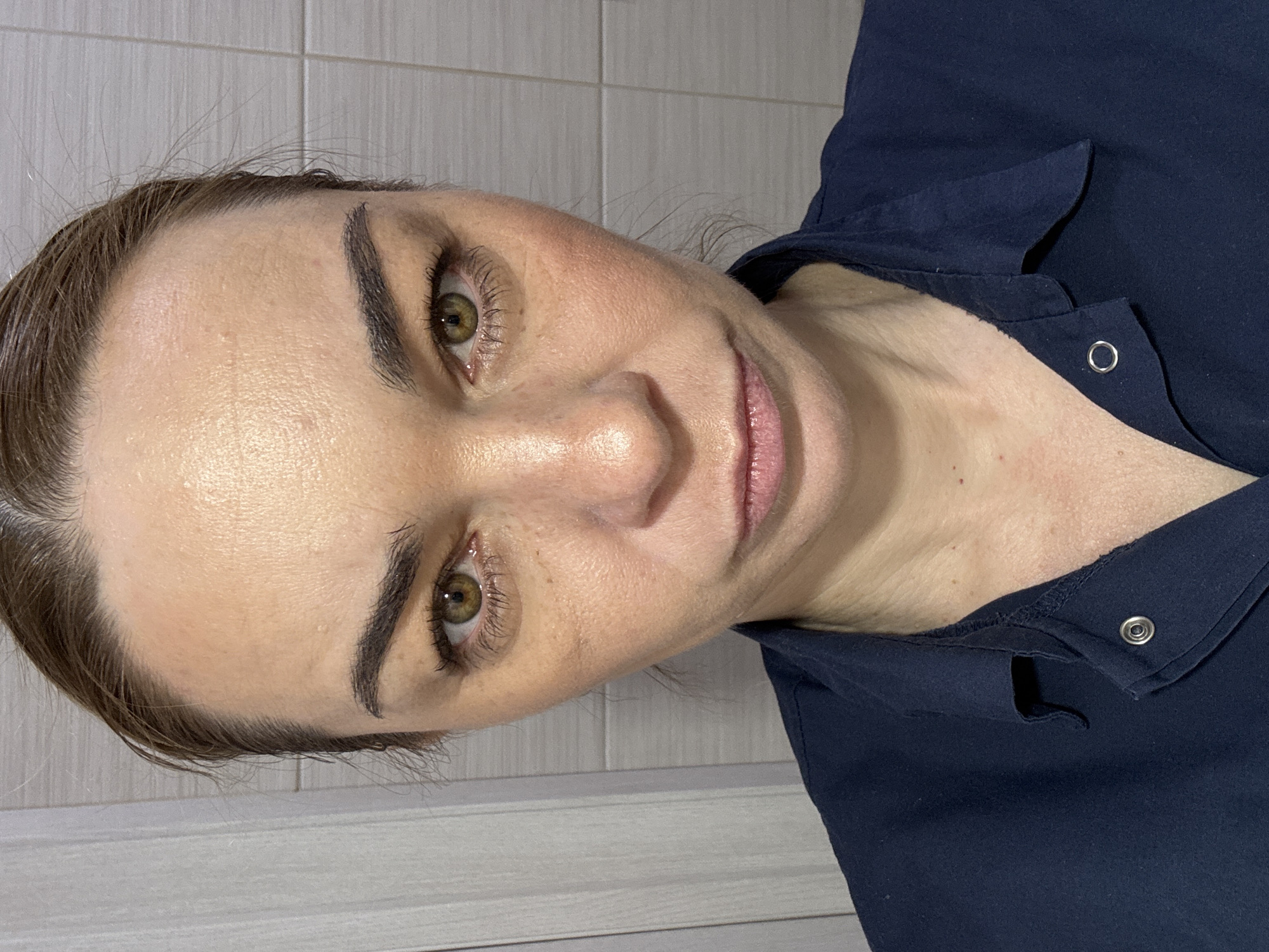 A woman with brown hair and green eyes is looking directly at the camera. She has fair skin and is wearing a dark blue collared shirt with metal snap buttons. The background consists of a light gray and white tiled wall. The image appears to be taken indoors, possibly in a bathroom or a room with similar tiled walls.