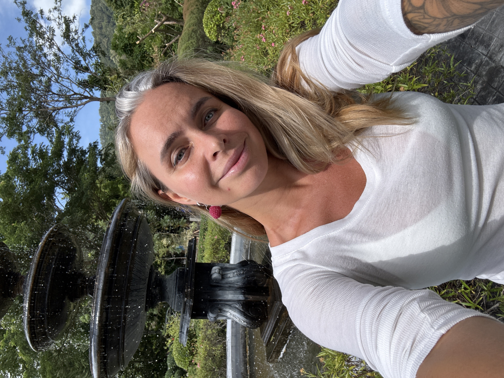 A woman with blonde hair and blue eyes is taking a selfie outdoors. She is wearing a white long-sleeved shirt. She has a pink earring on her left ear. Behind her, there is a multi-tiered fountain with water cascading down. Lush greenery, including trees and bushes, forms the background, with a clear blue sky and some clouds visible above. The scene suggests a park or garden setting. The image appears to have been taken in the district of Ko Samui, Thailand, characterized by its tropical vegetation and abundant water features.