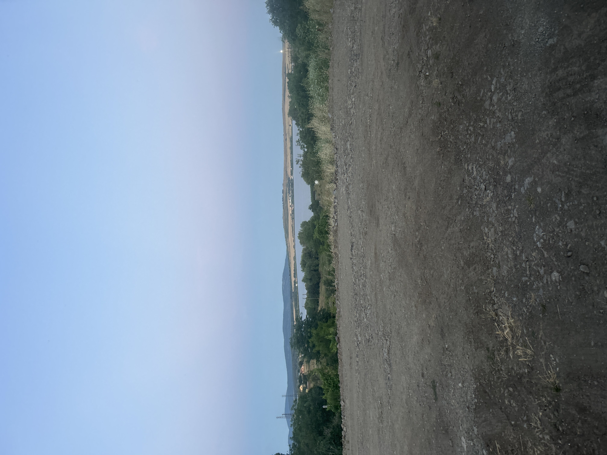 This image depicts a landscape scene with a body of water in the distance, likely a lake or bay, under a clear, pale blue sky. In the foreground, there is a dirt and gravel path, possibly leading away from the viewer. Beyond the path, a line of trees and vegetation separates the immediate terrain from the distant water. Hills or mountains are visible on the horizon beyond the water. The overall atmosphere suggests a natural or semi-natural environment, possibly on the outskirts of a populated area, given the presence of what appear to be some distant structures and power lines. The location is identified as being in Burgas, Bulgaria.
