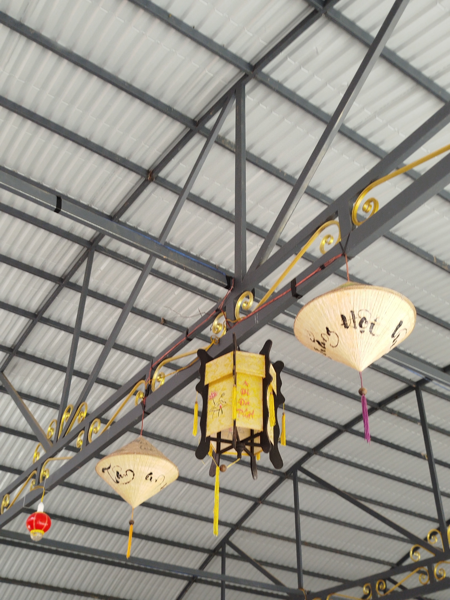 The photograph depicts an interior view, likely in Phát Chi, Vietnam, showing the underside of a corrugated metal roof supported by a complex steel frame. The steel frame is decorated with gold-colored ornamental swirls. Hanging from the frame are three distinctive light fixtures. Two appear to be conical hats with writing on them, one with a purple tassel and the other with a yellow tassel. The third light fixture is a box-shaped lantern with yellow panels and dark brown or black trim. Additionally, a small red lantern is visible hanging to the left, adding a festive or decorative element to the setting. The overall lighting is natural, casting shadows and highlighting the textures of the roof and the hanging elements.