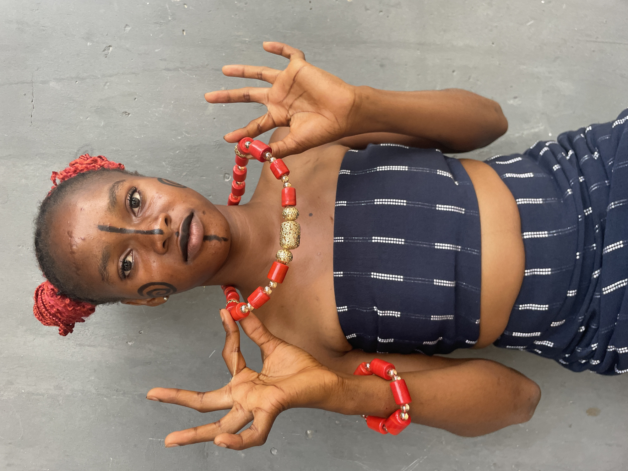 A young person with dark skin is lying on their back on a plain grey concrete floor, looking upwards and directly at the camera. The setting appears minimalist, suggesting an indoor studio or a simple outdoor area in Badagry, Nigeria.
The subject's face is adorned with intricate black body paint, featuring a bold horizontal line across the nose and cheeks, a distinct swirl pattern on their left cheek, and linear markings around their eyes and lips. Their hair is styled with vivid red braided accents visible on both sides of the head.
They are dressed in a dark blue, strapless garment, possibly a tube top or dress, patterned with horizontal white stripes. Around their neck is a prominent necklace crafted from large red beads interspersed with golden cylindrical and spherical beads. A matching red beaded bracelet is worn on their right wrist. The person holds the necklace with both hands, fingers spread in a stylized, expressive gesture, while maintaining a composed and direct gaze. The overall presentation suggests a cultural or traditional portrayal. There is no visible text.