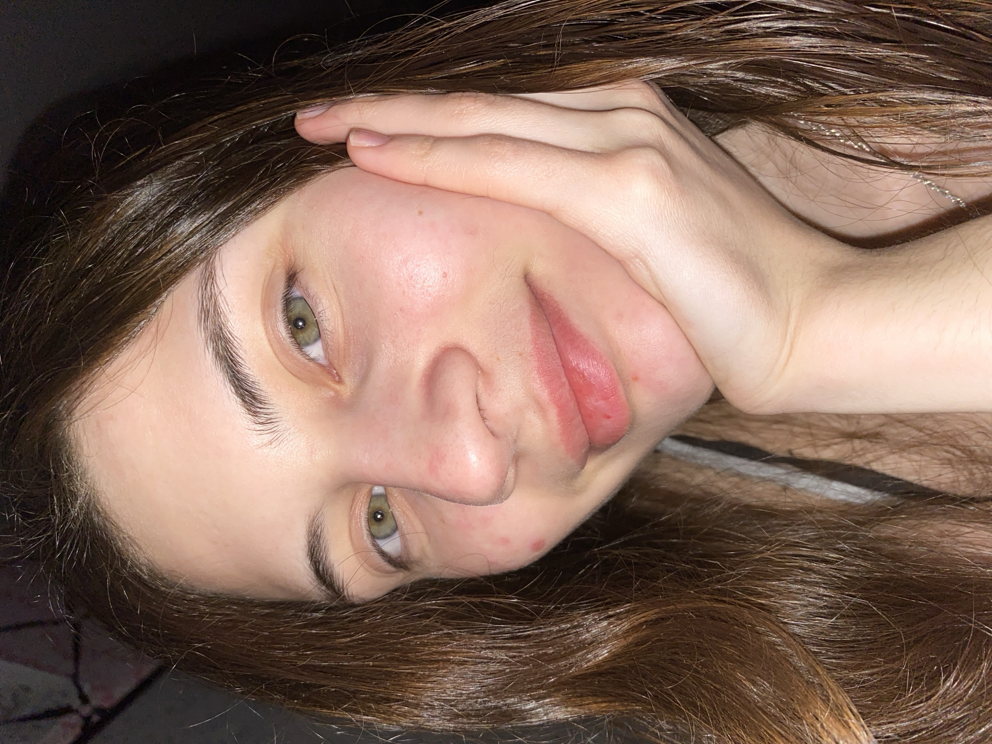 A close-up indoor portrait features a young person, likely female, with fair skin, long dark brown hair, and striking green eyes, taken in Votkinsk, Russia. The image is presented in a portrait orientation, with the subject's head tilted to the side.
The subject's face dominates the frame, with her gaze directed towards the viewer. One hand is casually placed on the side of her head, partially covered by her hair. Her long, wavy dark brown hair, which appears glossy, cascades around her face and shoulders, filling much of the background.
Visible details include minor redness and small blemishes on her cheeks and chin. Her lips appear full, with some visible redness or chapping. A thin, silvery necklace chain is subtly visible nestled within her hair on the right side of the frame. The background is indistinct and dark, suggesting an indoor setting with subdued or artificial lighting, likely taken during the evening or night. The subject's expression is neutral and direct, without a prominent smile. No text is visible in the image.