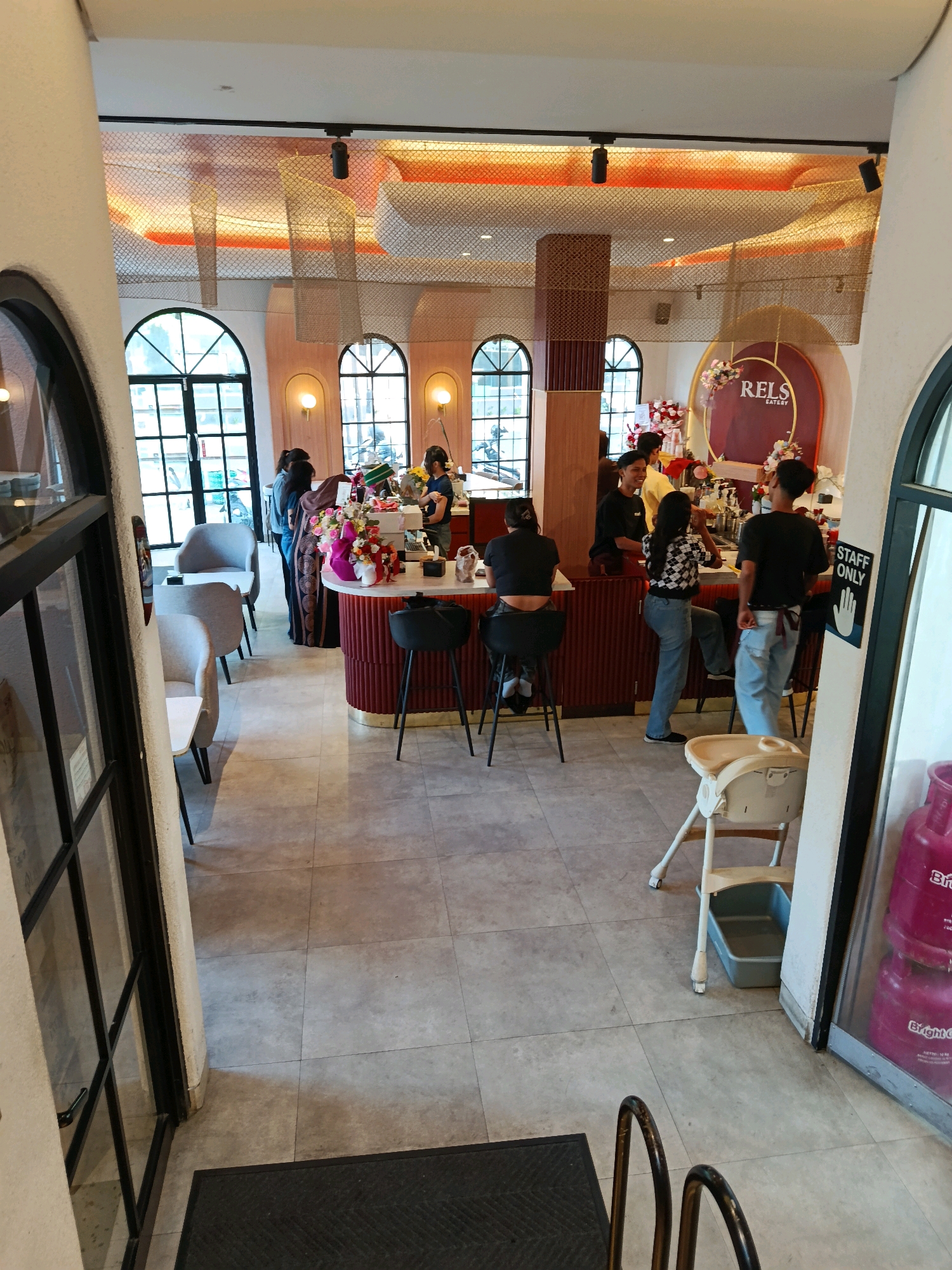 The image shows the interior of a cafe with a modern design. The cafe is located in Kudus, Indonesia. Several people are visible in the image. Two women are standing at the counter, and one woman is sitting on a stool. A male staff member is also visible behind the counter. The cafe has large windows, a bar area, and seating arrangements. The decor includes floral arrangements and a decorative logo on the wall. There are also some beverage dispensers visible in the background. The overall ambiance appears to be lively and contemporary.