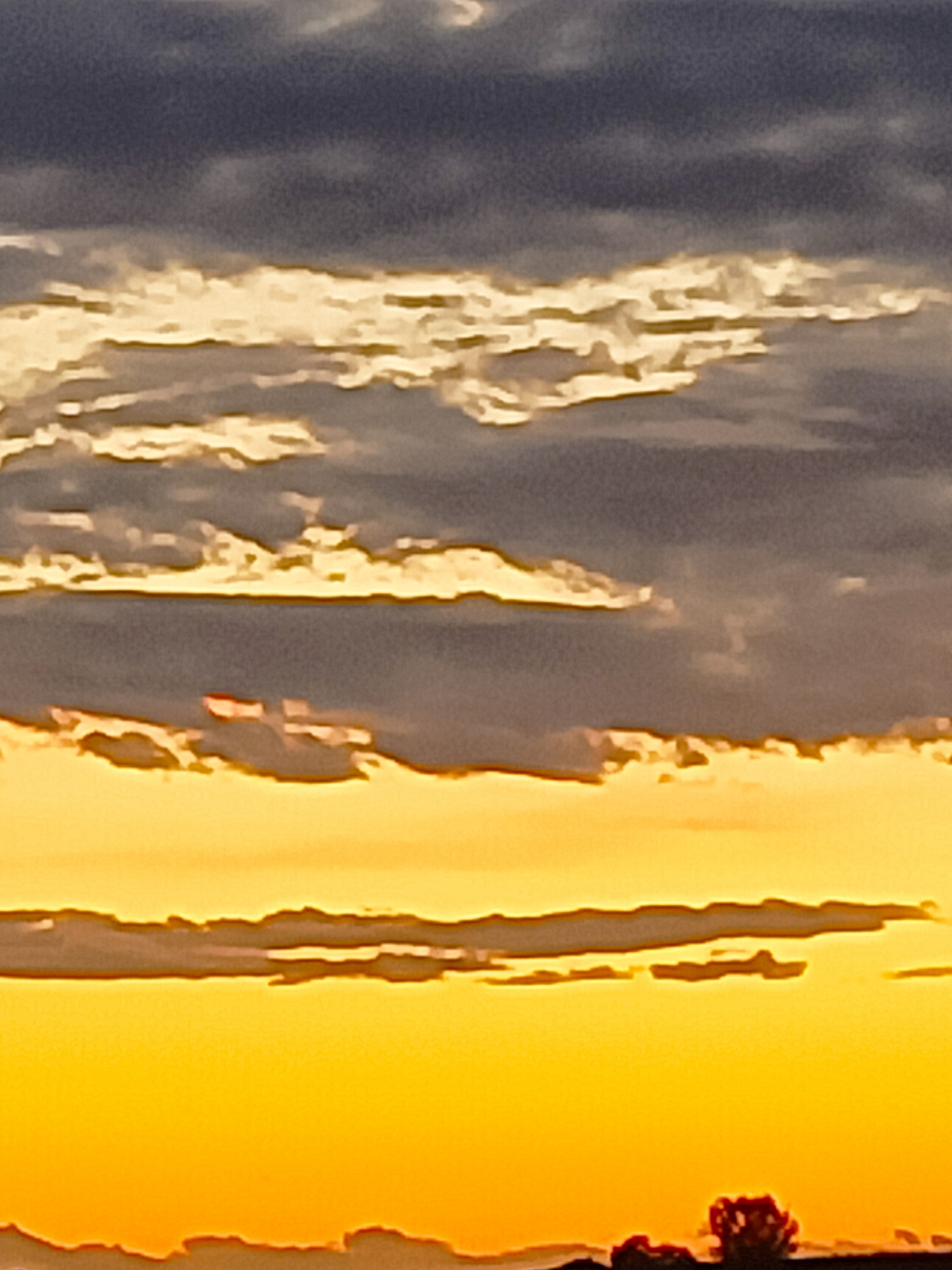 The image captures a sunset over a rural landscape. The sky is a gradient of warm colors, transitioning from a deep orange at the bottom to lighter yellow and then to gray and dark blue clouds at the top. Horizontal cloud formations are visible, some illuminated by the setting sun, creating streaks of bright yellow and white. In the foreground, a dark silhouette of the landscape is visible, with hints of trees and possibly low-lying structures. The image is abstract and lacks specific details of buildings or infrastructure that would definitively place it within Somaglia, Italy, but the style of the rural silhouette is consistent with the Italian countryside.
