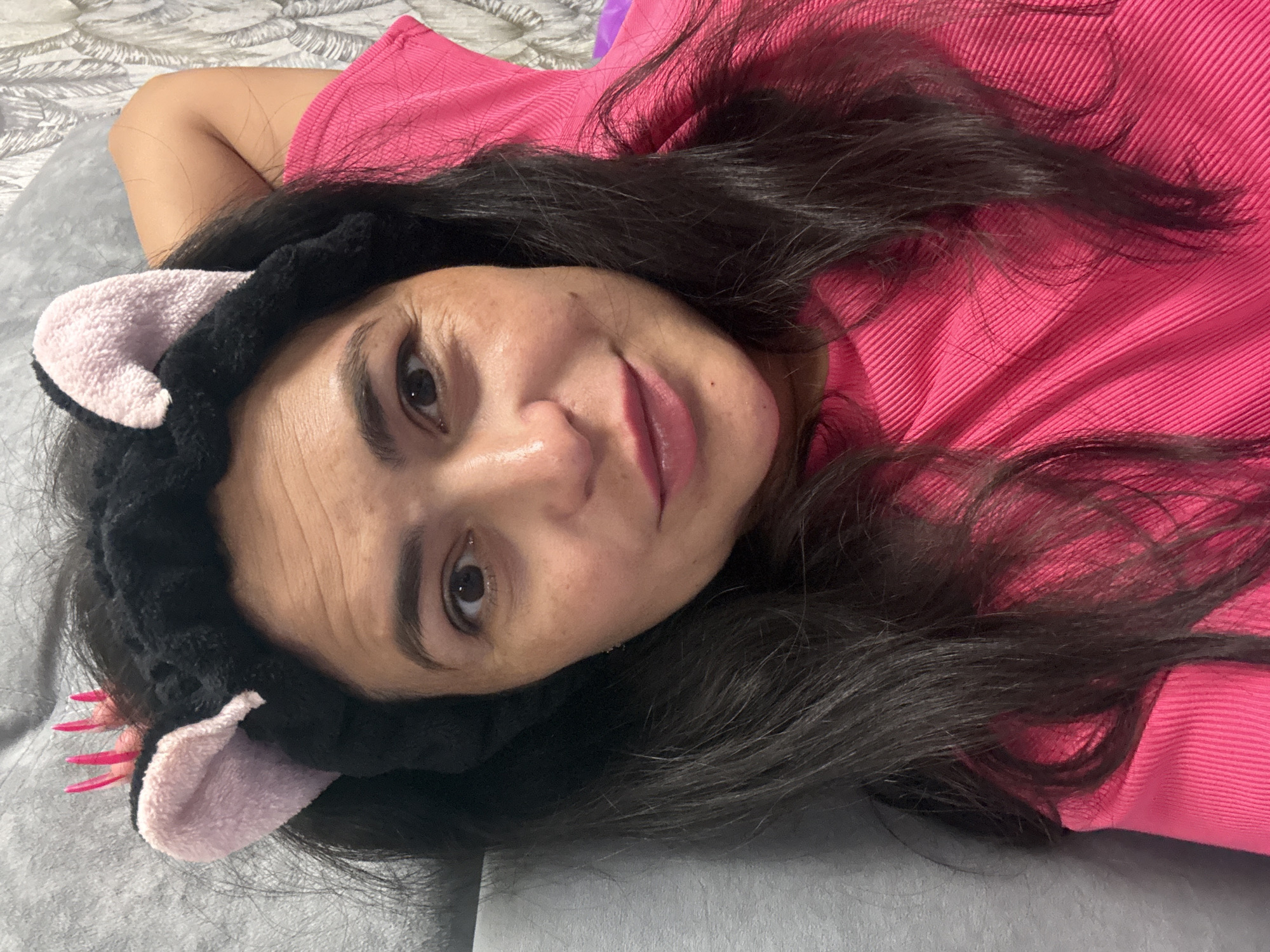 A person is lying on a gray surface, presumably a bed, and is wearing a pink shirt and a black headband with pink cat ears. The person has dark hair and is looking directly at the camera. A small purple object is visible in the upper portion of the image. There is no information to determine the location is Kauchuk Stantsiya, Russia.
