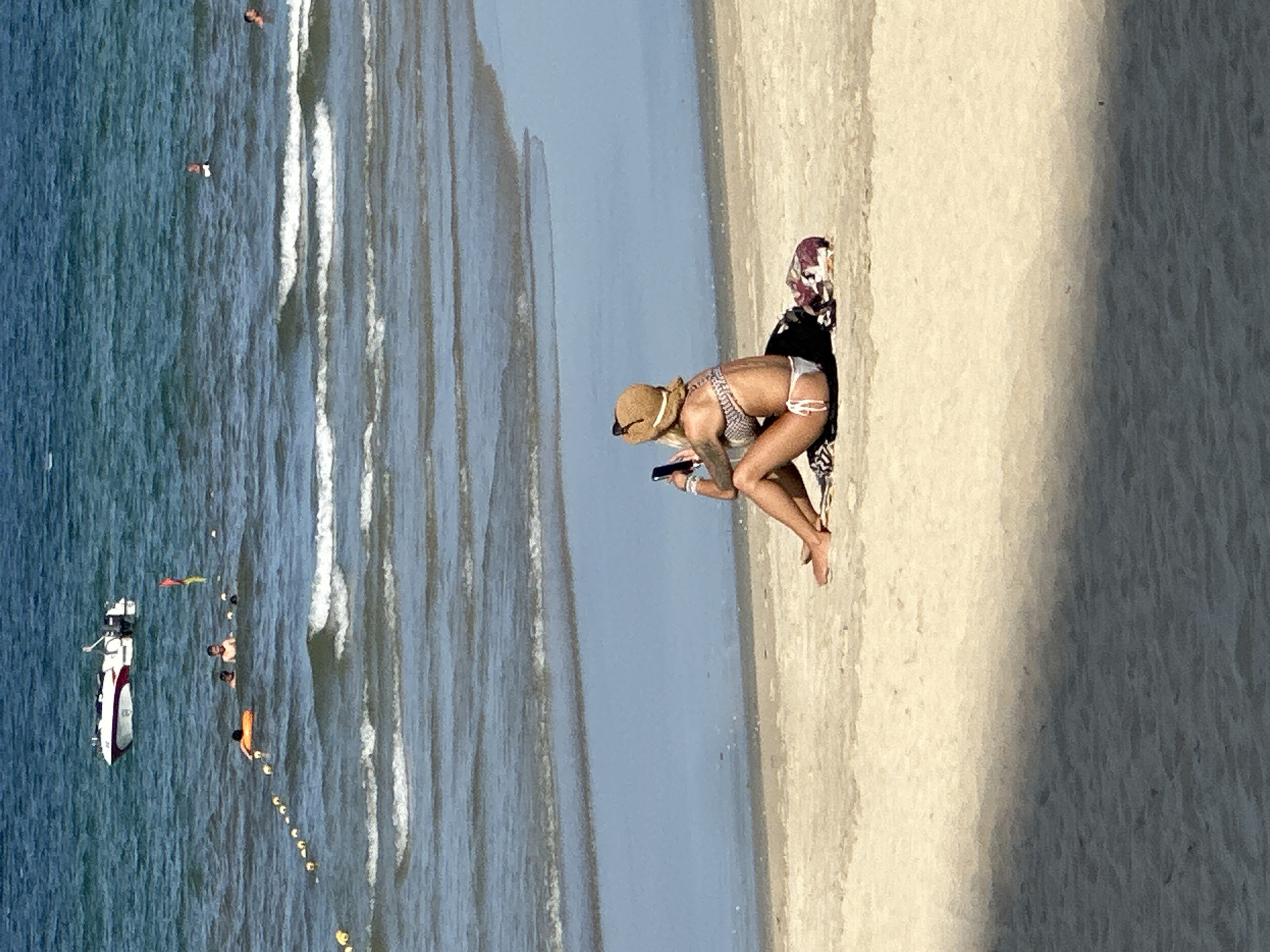 A woman wearing a bikini and a straw hat is sitting on a beach in Da Nang, Vietnam, looking at her phone. The beach is sandy, with waves crashing in the background. There are other people swimming in the ocean, and a boat is visible in the distance. The scene is bathed in sunlight, with a shadow cast across the sand in the foreground.