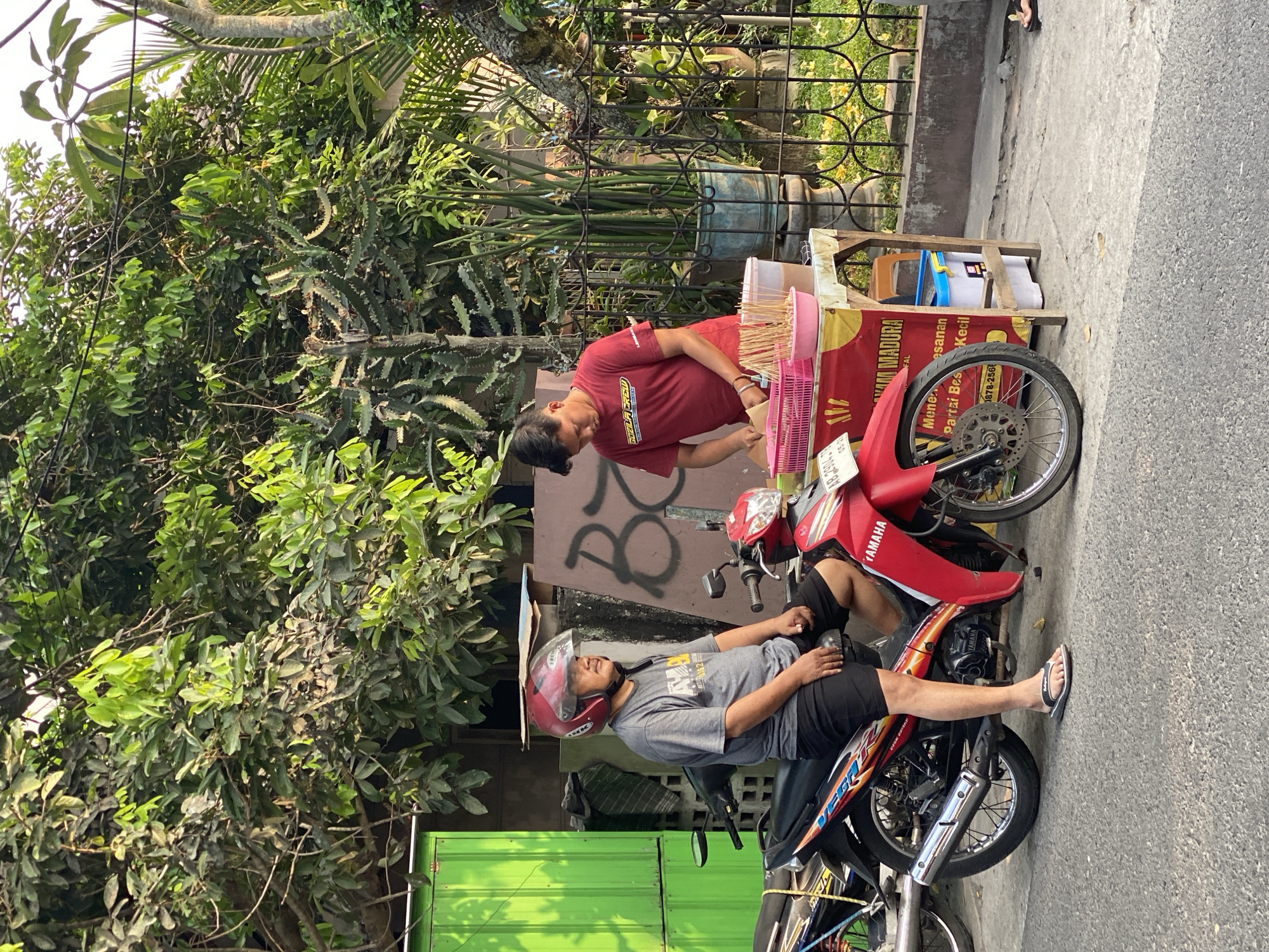 In Kasihan, Indonesia, two men are visible. One man, wearing a red helmet, a grey t-shirt, and black shorts, sits on a red motorcycle parked on an asphalt road. He appears to be waiting. The second man, in a red t-shirt, stands behind a small street food cart, preparing skewers. The cart displays text indicating "AYAM MADURA" and "Mene... esanan Partai Bes... Kecil." Behind the men and the cart, there is lush green foliage, including trees and a cactus plant. A green metal structure and a wall with graffiti are also present in the background. A black metal fence encloses a garden area with yellow flowers. The scene appears to be a street-side food stall during daylight.