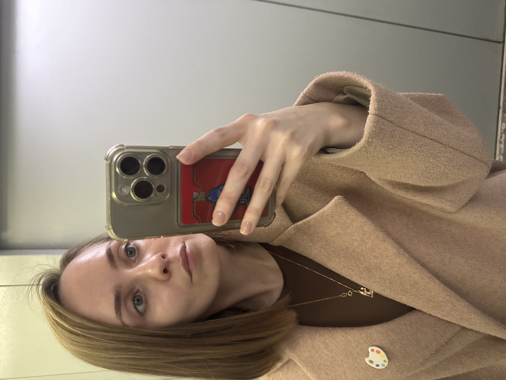 A young adult female is captured in a mirror selfie, visible from the upper torso, within an elevator interior in Perm, Russia. She has light brown, shoulder-length hair, styled straight and parted on her right side, and light blue-green eyes. Her complexion is fair. She wears a dark brown top layered under a textured beige wool-blend coat. A small, white art palette pin, detailed with red, orange, yellow, green, light blue, and purple dots, is fastened to her left lapel. A fine gold-colored necklace features a small, stylized gold figure pendant. Her right hand, displaying nails with nude polish, holds a smartphone, likely an iPhone Pro Max model, encased in clear material, revealing three camera lenses. A red magnetic wallet or cardholder, embossed with a blue and white figure, is attached to the back of the phone case. The background consists of smooth, light-colored vertical panels, consistent with elevator walls, under bright, uniform lighting.