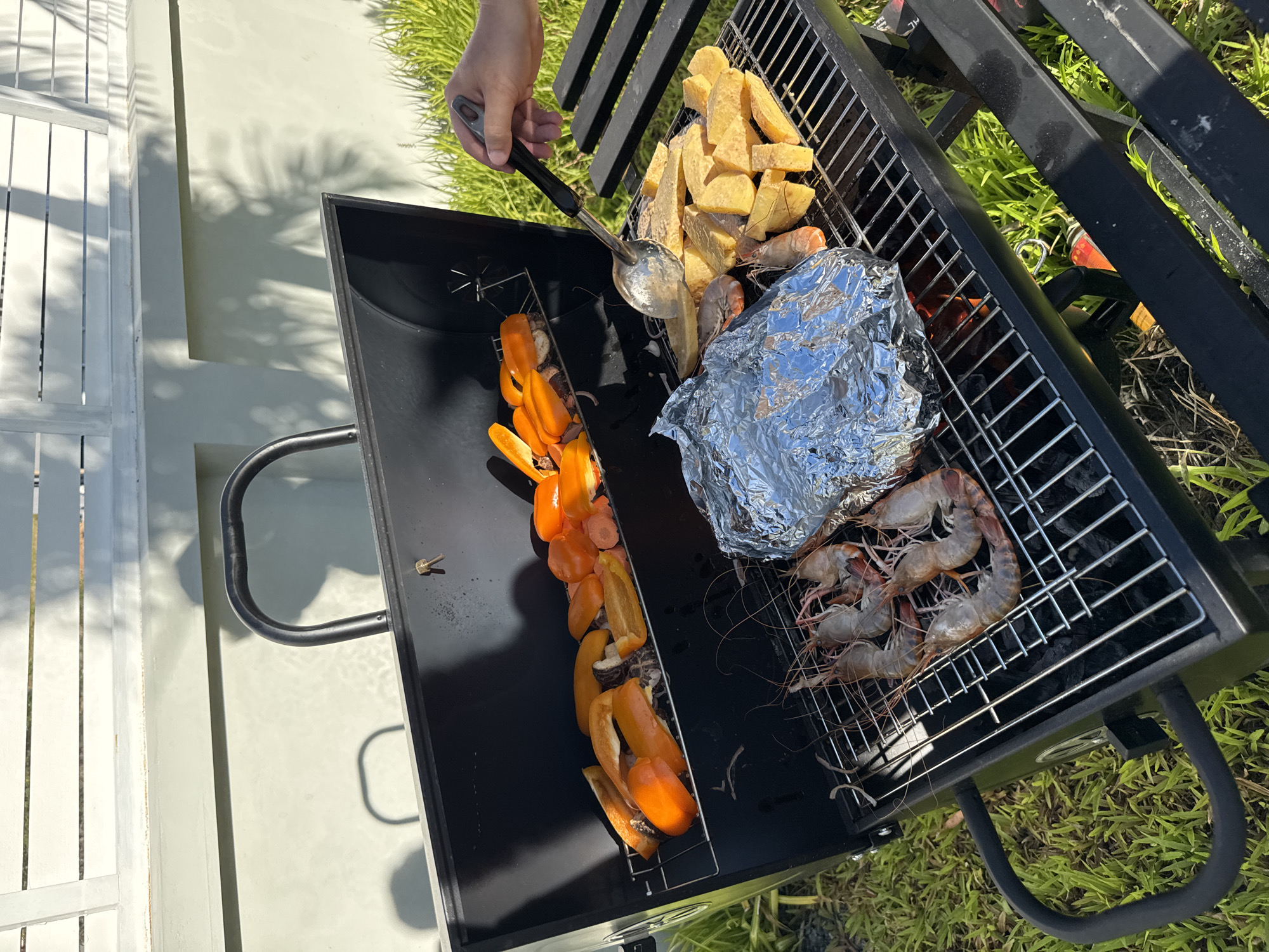 The image shows a close-up, overhead view of a black charcoal grill set up outdoors on a patch of green grass. The scene suggests a sunny day, with bright natural light and distinct shadows cast on a light-colored wall and white slatted fence in the background. The setting appears to be a backyard or garden area, possibly in Baan Tai, Thailand, given the lush green vegetation.
On the grill's grates, several food items are being prepared. To the left, a metal rack holds several pieces of chopped orange bell peppers and some carrots, skewered together. In the center, a pile of potato wedges is situated on the grates, and next to them, a packet of food wrapped in aluminum foil. To the right, a cluster of large, raw shrimp with visible antennae are arranged on the grill. The grill itself has a dark, potentially heat-resistant surface and black metal legs, with a black handle visible on the left side.
A person's hand is visible in the upper left quadrant of the image, holding a metal ladle. The hand is in the process of scooping something from the grill, though the exact item being scooped is unclear. The overall mood conveyed is one of casual outdoor cooking and preparation for a meal. There is no visible text in the image.