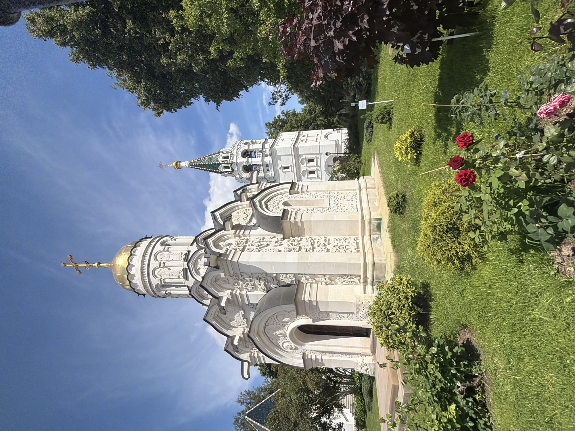 The image displays a picturesque architectural complex, likely an Orthodox Christian church and bell tower, nestled within a lush garden setting. The main building is a white, ornate church, characterized by a prominent golden dome topped with an Orthodox cross. Its facade is richly decorated with intricate carvings. Behind it, and slightly to the right, stands a taller, slender bell tower, also white, with a distinctive green and white patterned spire, similarly crowned by a golden cross.
The environment is a well-maintained garden featuring expansive green lawns, various bushes (some with dark red foliage), and tall green trees framing the scene from the top and sides. In the foreground, vibrant red and pink roses bloom on bushes. The weather appears bright and sunny, with a clear blue sky dotted with a few scattered white clouds. The time of day is clearly daylight. No people are visible, contributing to a tranquil and serene atmosphere. Small white signs on poles are present in the grassy areas, but their text is illegible. The architectural style and context suggest this scene is located in Pavlovskaya Sloboda, Russia.