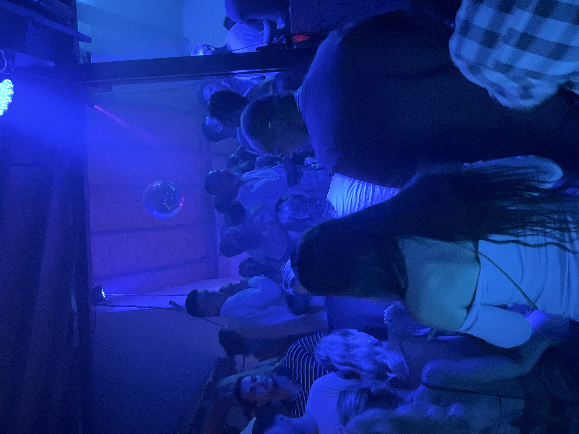 The image depicts a crowded indoor venue, likely a nightclub or bar, illuminated by vibrant blue and purple lights. Several individuals are visible, with some in focus and others blurred due to movement or distance. The atmosphere appears to be lively, with a disco ball hanging from the ceiling, reflecting the lights. In the foreground, a person is seen from behind, wearing a dark top. Other individuals in the midground and background are also engaged in social interaction or dancing. The architectural elements of the venue, such as exposed wooden beams and ceiling structures, are partially visible. The overall scene suggests a nighttime social gathering. No specific location in Herceg Novi, Montenegro, can be determined from the image alone.