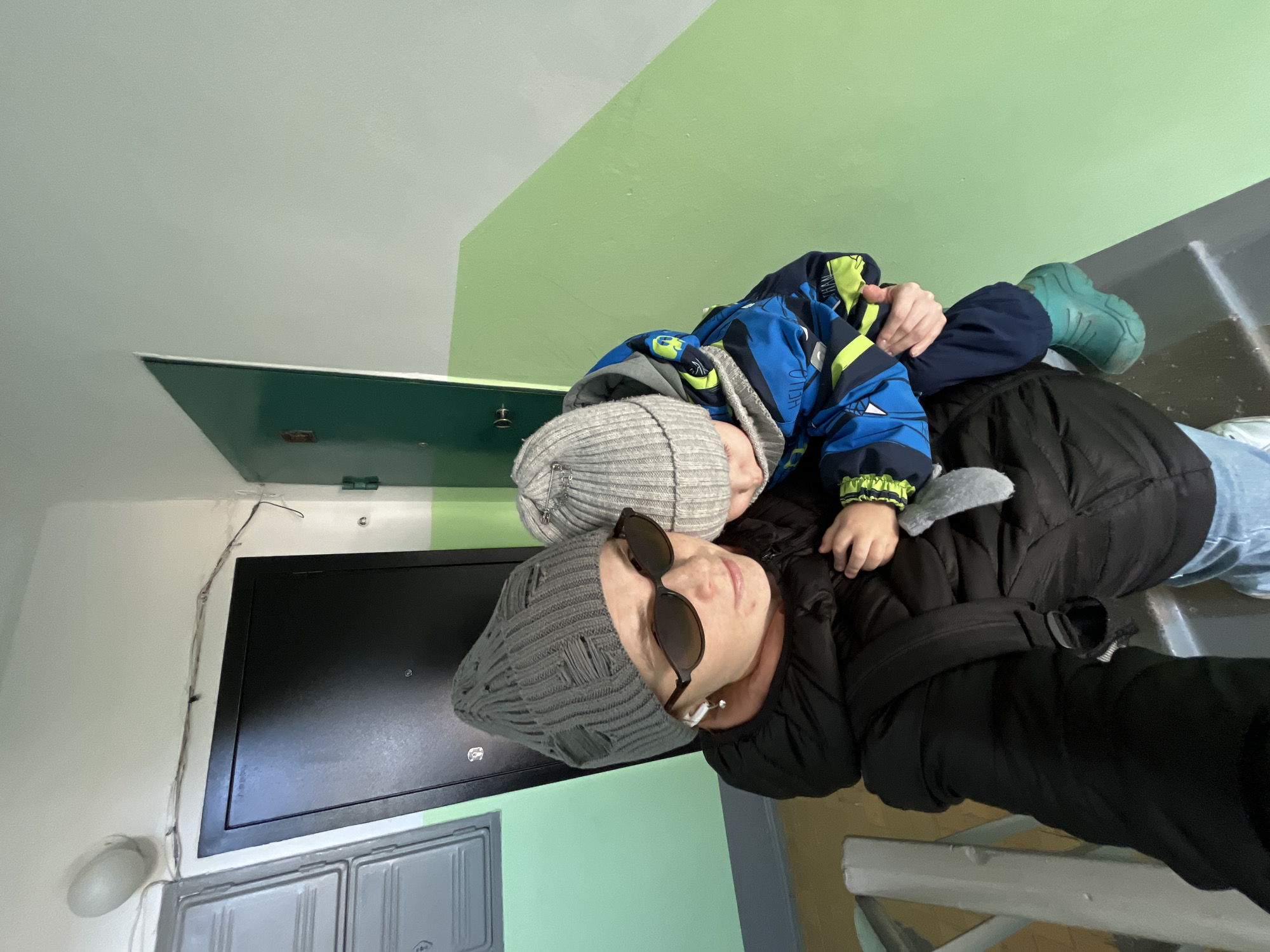 A woman wearing a grey beanie and sunglasses is lying down on what appears to be a landing of stairs inside a building, with a young child wearing a grey beanie, blue jacket, and green boots lying on top of her. The woman is wearing a black puffy jacket and light blue jeans. The landing walls are painted in a two-tone scheme of white and green, with a black metal door visible in the background. A metal staircase with white railings is in the lower right corner. The photograph was most likely taken inside an apartment building in Chaikovsky, Russia.