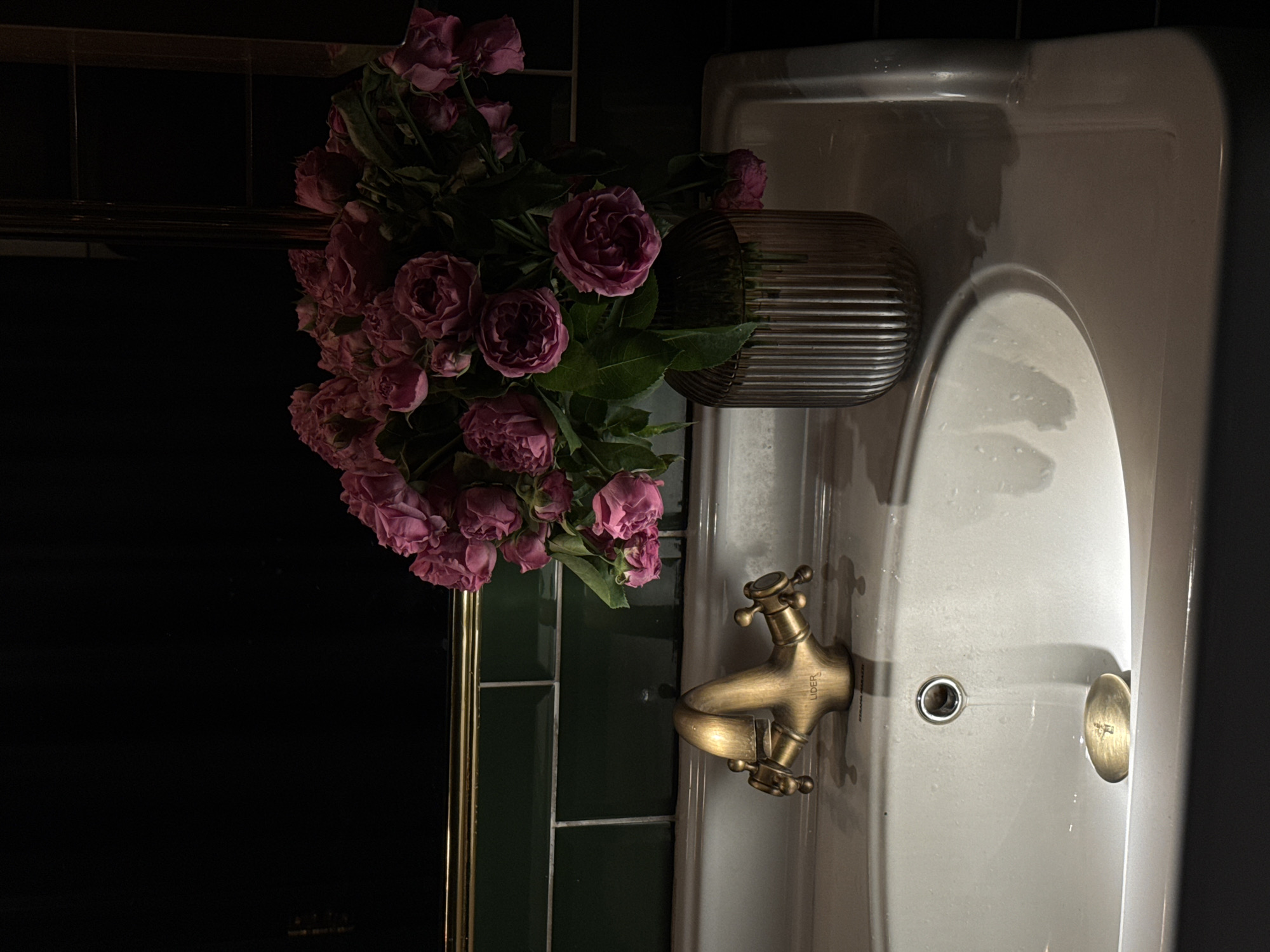 The image shows a bathroom scene, with a white porcelain sink and a bronze-colored faucet. A vase of pink roses is positioned to the left of the sink. The wall behind the sink is tiled with dark green rectangular tiles. To the left of the sink is a dark, possibly mirrored surface with a gold frame. The overall lighting is dim, creating a moody atmosphere. The location is potentially Yerevan, Armenia, but there is no explicit information in the image to confirm this.