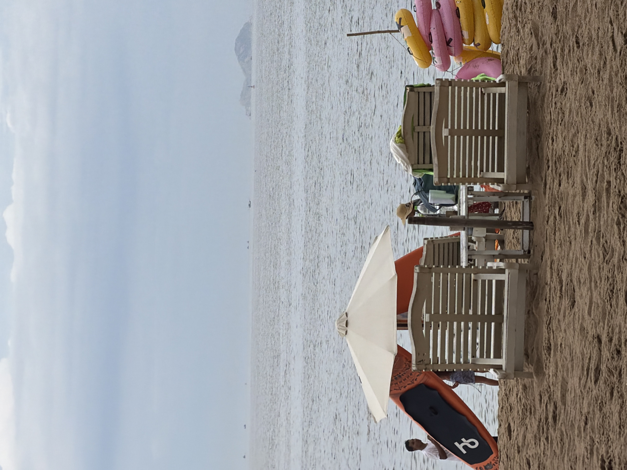 The image depicts a cloudy day at a beach in Nha Trang, Vietnam. In the foreground, the sand is a warm brown, with subtle footprints. To the left, a man in a white shirt stands near the water's edge, holding an orange paddleboard with a black section. Behind him, a portion of a beach umbrella is visible. In the middle ground, two light-colored wooden lounge chairs are set up on the sand, facing the ocean. A small white table is positioned between them. A person wearing a hat and a blue skirt is seated between the chairs. To the right, a stack of colorful inflatable rings, predominantly pink and yellow, leans against a thin wooden pole. In the background, the sea stretches out to the horizon, with a few distant islands visible under a hazy sky. Several small boats or distant objects are scattered across the water. The overall scene suggests a calm, overcast day at the beach.