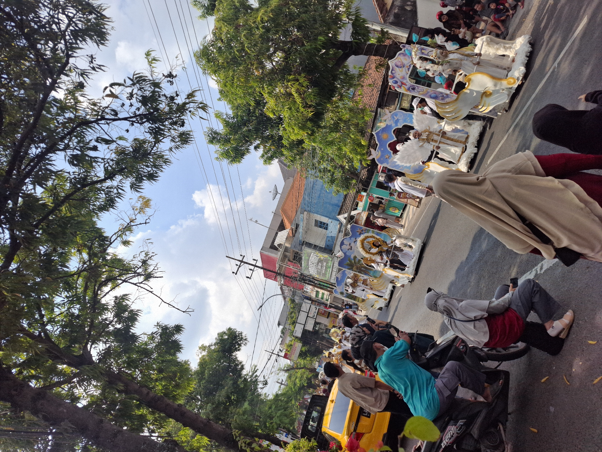 A bright, sunny day in Lamongan, Indonesia, is depicted in this image, with a parade passing by on a street lined with trees and buildings. The parade features elaborately decorated floats, with the most prominent ones designed as fantastical chariots. Several children, dressed in white costumes and elaborate headdresses, are positioned on these floats. One child appears to be dressed as an angel, with wings. The floats are adorned with white, cloud-like material and golden accents, with some featuring celestial motifs like suns and moons.
On the street, several people are watching the parade. A yellow car is visible on the left side of the image. Two individuals are seated on a motorcycle, with the rider wearing a turquoise shirt and the passenger in a grey hijab and red shirt. Another person in a grey hijab and red skirt is also visible near the motorcycle. Spectators are gathered on the right side of the street, with some sitting down to get a better view. The buildings in the background suggest an urban setting, with a mix of traditional and modern architecture. Power lines crisscross the sky, and trees with lush green foliage provide shade over the scene. The overall atmosphere appears festive and celebratory.