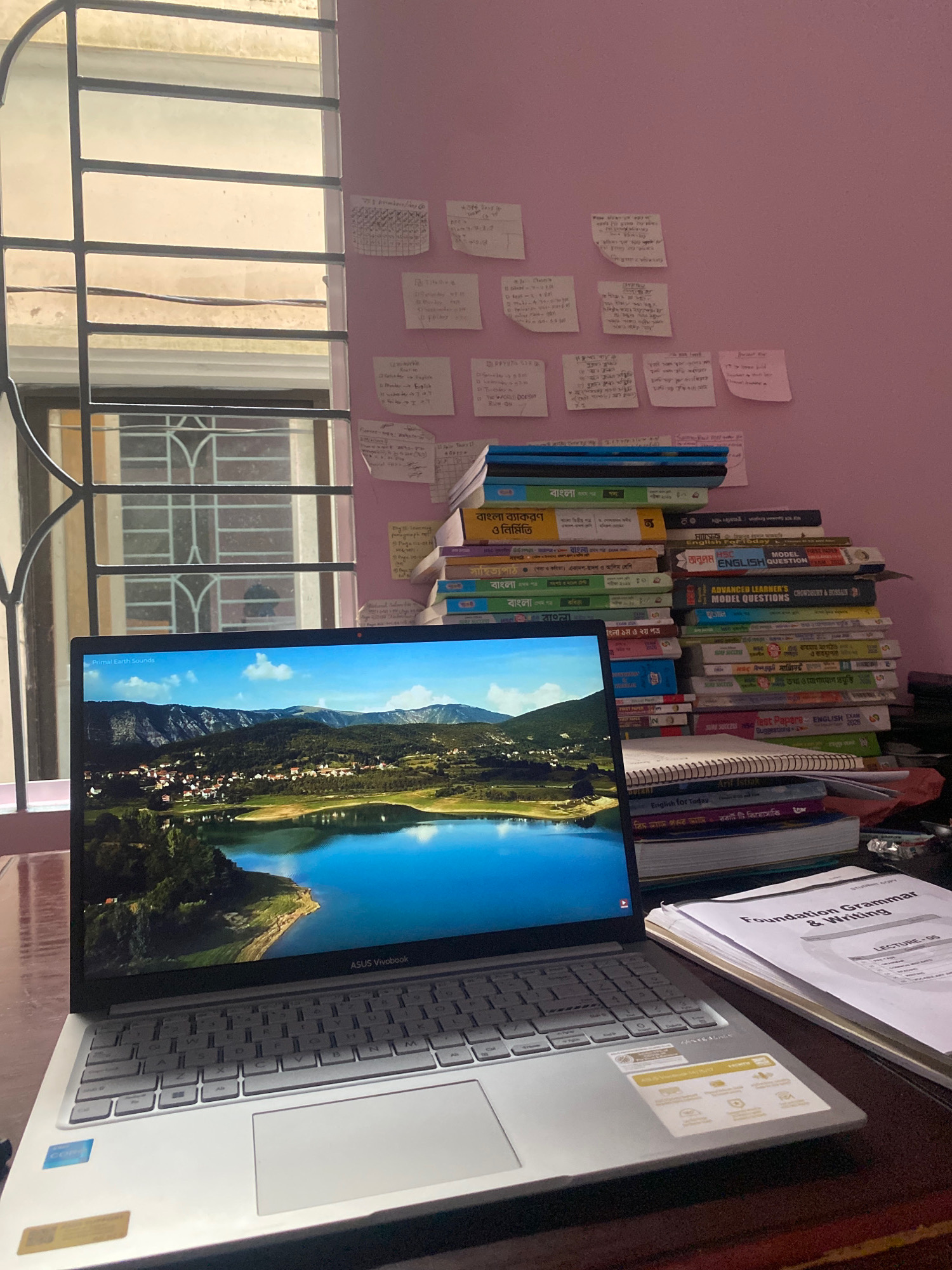The image depicts a desk in a room, likely in Hemayetpur, Bangladesh, with a window providing natural light. A silver ASUS Vivobook laptop is open, displaying a scenic landscape on its screen. To the right of the laptop, a stack of books and notebooks is piled high, some with Bengali text on their covers, suggesting academic or study materials. A document titled "Foundation Grammar & Writing" is placed on top of another notebook. The wall behind the desk is painted pink and is covered with numerous sticky notes, some containing handwritten text. The overall scene suggests a dedicated study or work environment.
