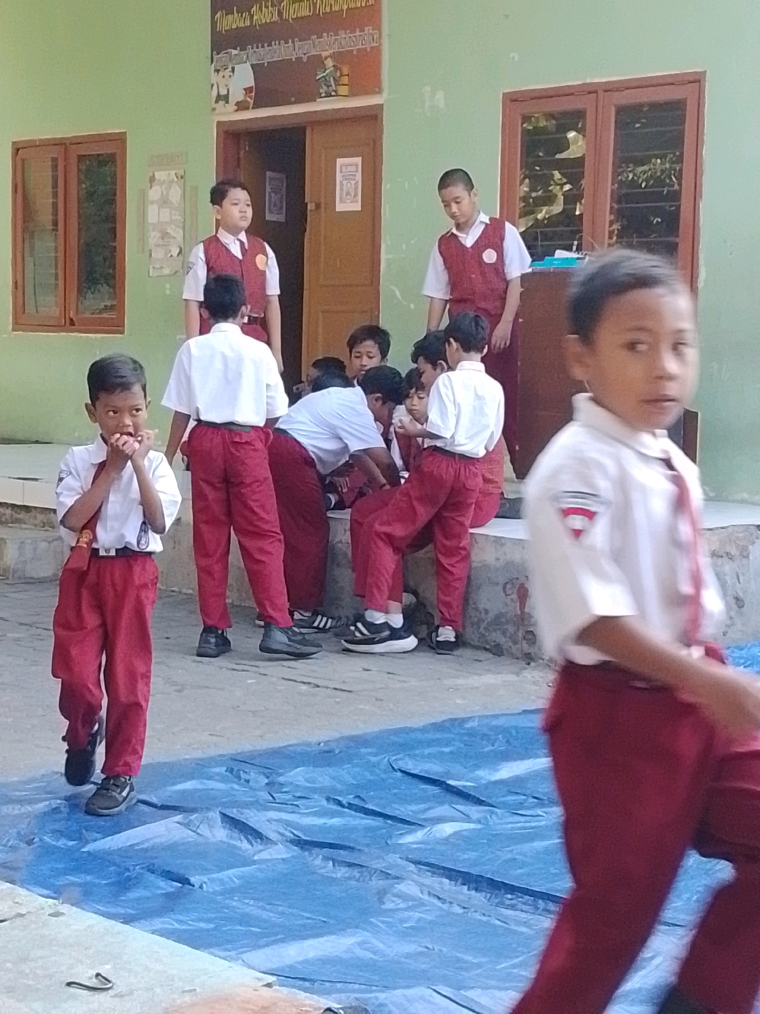 In the city of Surabaya, Indonesia, a group of young male students, likely of elementary school age, are gathered outside a building with a light green facade. They are all wearing similar school uniforms consisting of a white short-sleeved shirt and red trousers, with some also wearing red vests. The students are engaged in various activities, with some standing, others sitting, and one in the foreground appearing to put something in his mouth. The environment suggests a school setting, with a window and a doorway visible on the building. A large blue tarp covers the ground in front of the students. The overall scene depicts a casual moment among schoolchildren in an outdoor educational setting.