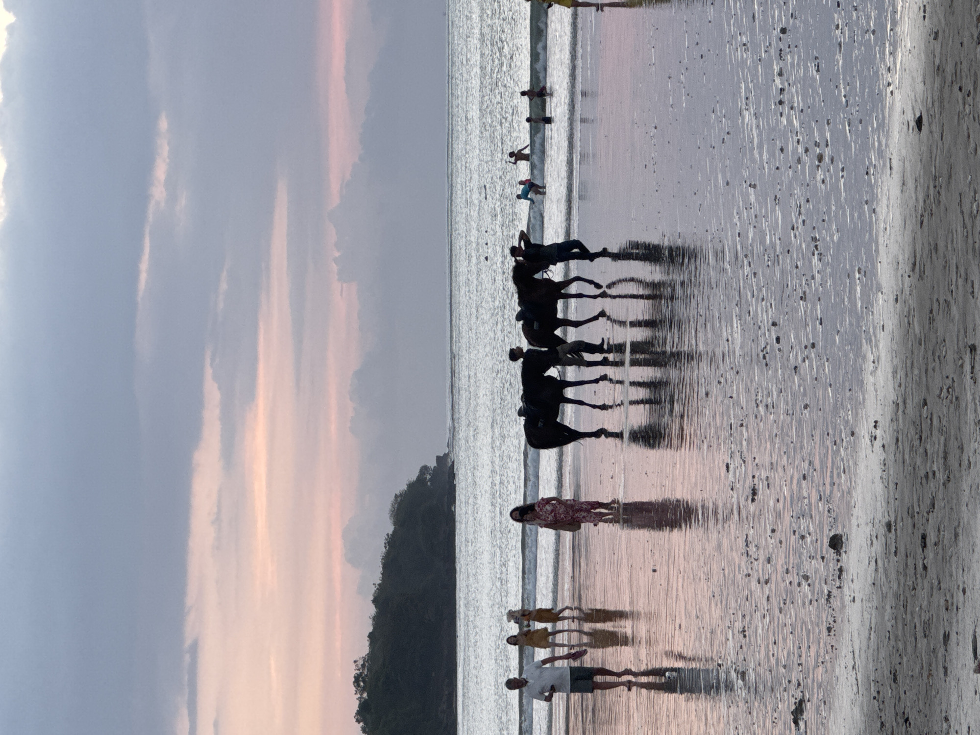 The image captures a picturesque beach scene at what appears to be sunset or sunrise in Kuta Selatan, Indonesia. The sky above is a blend of soft gray clouds and vibrant pastel hues of pink, orange, and purple, indicating either dawn or dusk.
The foreground and mid-ground are dominated by the wide expanse of wet sand, which beautifully reflects the colors of the sky, the figures on the beach, and the distant landmass. In the mid-ground, two dark-silhouetted horses, each carrying a rider, are walking along the water's edge, their long, distinct shadows stretching across the reflective sand. Further out in the ocean, several people are seen wading or swimming in the gentle, breaking waves. Closer to the viewer, a couple stands on the wet sand, their forms also mirrored by the reflective surface, along with other individuals scattered along the shoreline.
To the left, a dark, densely forested hill or landmass rises, providing a dramatic backdrop to the beach. The ocean exhibits moderate waves near the shore, with the foamy white crests contrasting with the smooth, mirror-like sand. The scene conveys a sense of tranquility and leisure, highlighting the natural beauty of the coastal environment during the golden hour. There is no visible text in the image.
