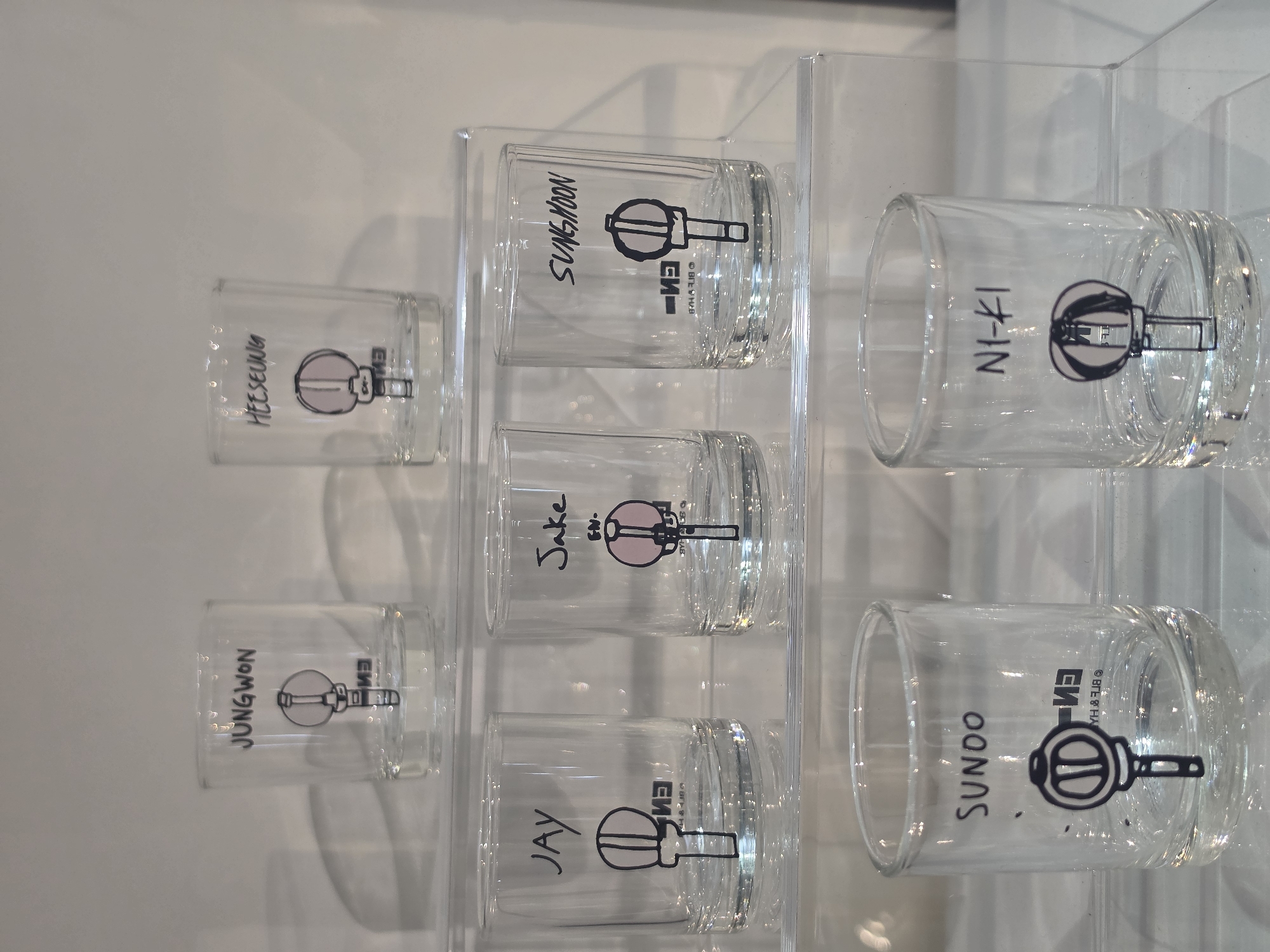 The image displays a collection of seven clear glass cups or shot glasses, arranged neatly on a transparent acrylic or glass display shelf against a plain light-colored wall. The setting appears to be indoors in Bangkok, Thailand, characterized by bright, even lighting.
Each glass features a vertically printed name in black lettering, along with a stylized graphic resembling a lightstick or lollipop, and the text "EN-" or "EN". The names visible on the glasses are HEESEUNG, JUNGWON, JAY, Jake, SUNGHOON, SUNOO (mistranscribed as SUNDO in OCR), and NI-KI. These names correspond to the members of the K-pop group ENHYPEN, and the "EN-" text likely refers to the group.
The glasses are organized in two rows: three on the left stack (HEESEUNG, JUNGWON, JAY) and four on the right stack (Jake, SUNGHOON, SUNOO, NI-KI), though Jake and SUNGHOON are in the upper part of the right stack, and SUNOO and NI-KI are in the lower part. The small lightstick/lollipop graphic next to some names, like HEESEUNG and Jake, has a pink tint, while others are monochrome. One cup (SUNOO) also shows mirrored text that appears to be "© BIGHIT", indicating the copyright holder. No specific activity is taking place, as the image captures a static display of merchandise.