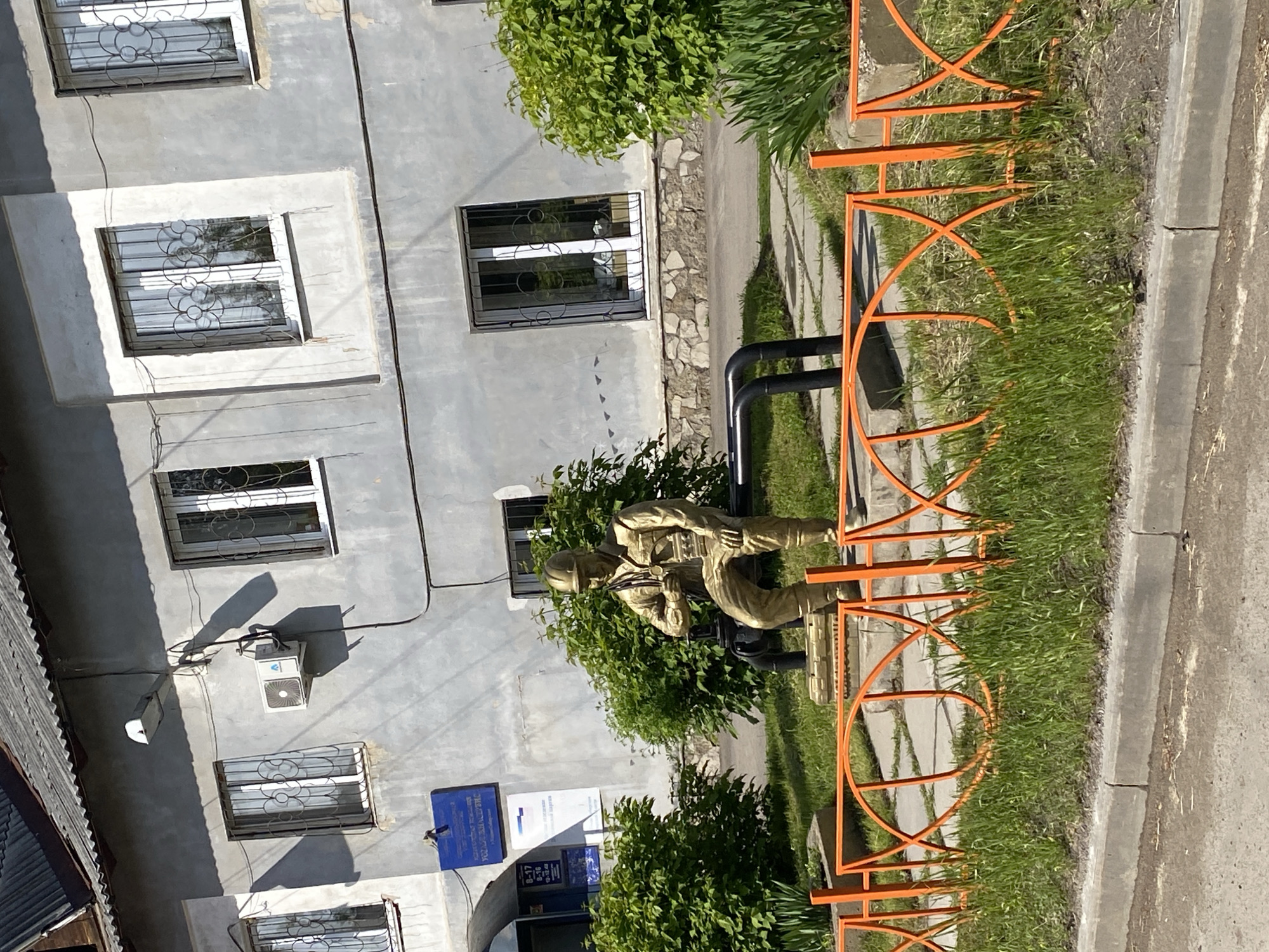 This image captures a street scene in Usolye-Sibirskoye, Russia, on a sunny day, likely mid-day or early afternoon, as evidenced by the distinct shadows.
The main subject is a multi-story building with a light gray or off-white stucco facade. It features multiple rectangular windows, each secured with decorative black metal grates. An air conditioning unit is mounted on the wall of the building, and various electrical cables run along its exterior. On the lower part of the building, a prominent blue sign is visible, displaying text in Cyrillic. The most clearly discernible word on the sign is "ПОЛИЦИЯ" (Police), indicating the building is likely a police station or a related administrative office. Additional text below suggests office hours or departmental information.
In front of the building, nestled within green bushes, stands a golden-tan statue of a person. The figure, dressed in what appears to be a uniform and helmet, is depicted in a crouching or kneeling position, holding what resembles a rifle or similar equipment. The statue adds a commemorative or thematic element to the scene.
An orange metal fence with decorative, intertwined circular patterns separates the building's immediate surroundings from the street. This fence runs alongside a paved area, a strip of green grass, a concrete curb, and an asphalt road. The setting combines elements of urban architecture with natural greenery and public infrastructure.