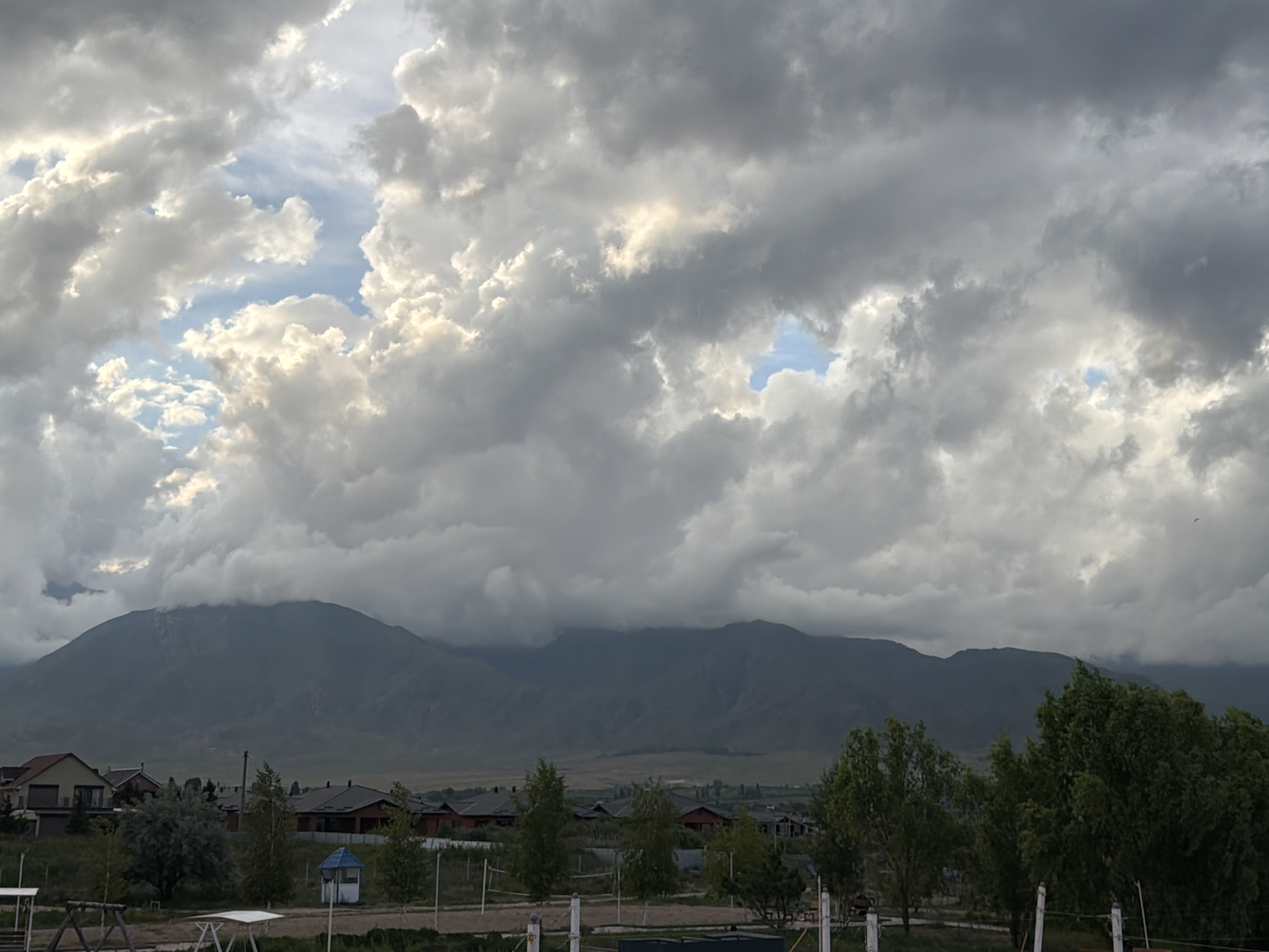 The image depicts a wide landscape under a dramatic sky filled with heavy, textured clouds. Patches of bright blue sky are visible through breaks in the clouds, illuminated by sunlight. Below the sky, a range of dark, imposing mountains dominates the horizon, their peaks partially obscured by low-hanging clouds.
In the foreground, a settlement of houses with red-tiled roofs is nestled at the foot of the mountains. Trees, some with lush green foliage and others appearing more bare, are scattered throughout the scene, adding depth and texture. In the immediate foreground, what appears to be a playground structure and fencing suggests a residential or recreational area. A small, blue-roofed structure, possibly a shelter or utility box, is also visible.
The time of day appears to be late afternoon or early evening, given the dramatic lighting and cloud formations. The weather seems to be changing, with a mix of sunlight breaking through dark clouds, suggesting a possibility of rain or an approaching storm. The location cues, such as the mountain range and the architectural style of the houses, are consistent with the provided context of Çolpon-Ata, Kyrgyzstan. The overall mood is one of natural grandeur and atmospheric intensity. No text is visible in the image.