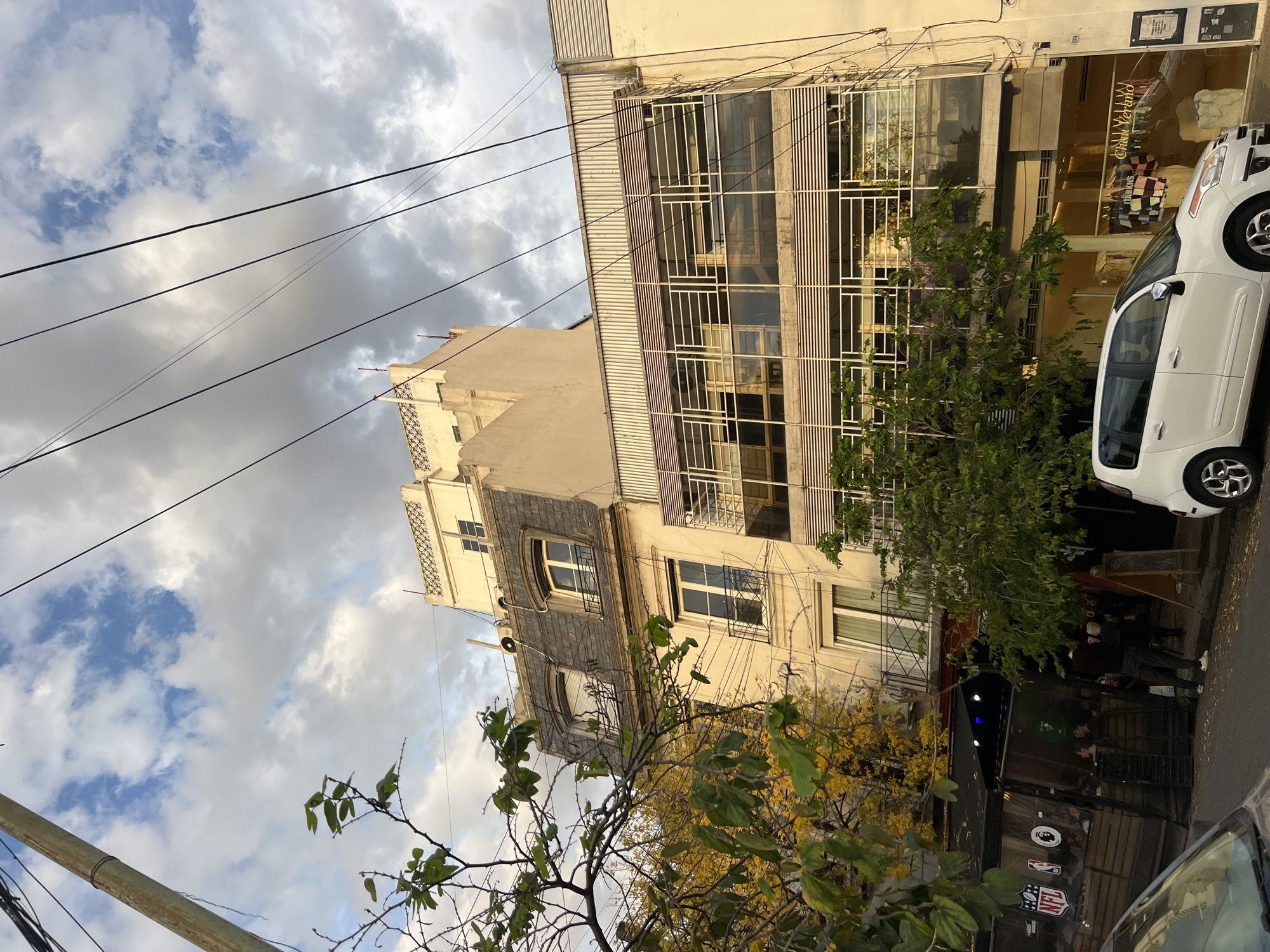 The image shows an outdoor scene in Buenos Aires, Argentina, with a mix of architectural styles under a partly cloudy sky. In the foreground, there's a car parked along the street. A building with a mix of modern and classic design elements is visible, featuring large window grids and balconies. The street-level area seems to have a storefront or restaurant with people present. Above, there's a classic apartment building with ornate facades and a sloped roof. Utility wires stretch across the sky. Trees with yellowing leaves partially obscure the view.