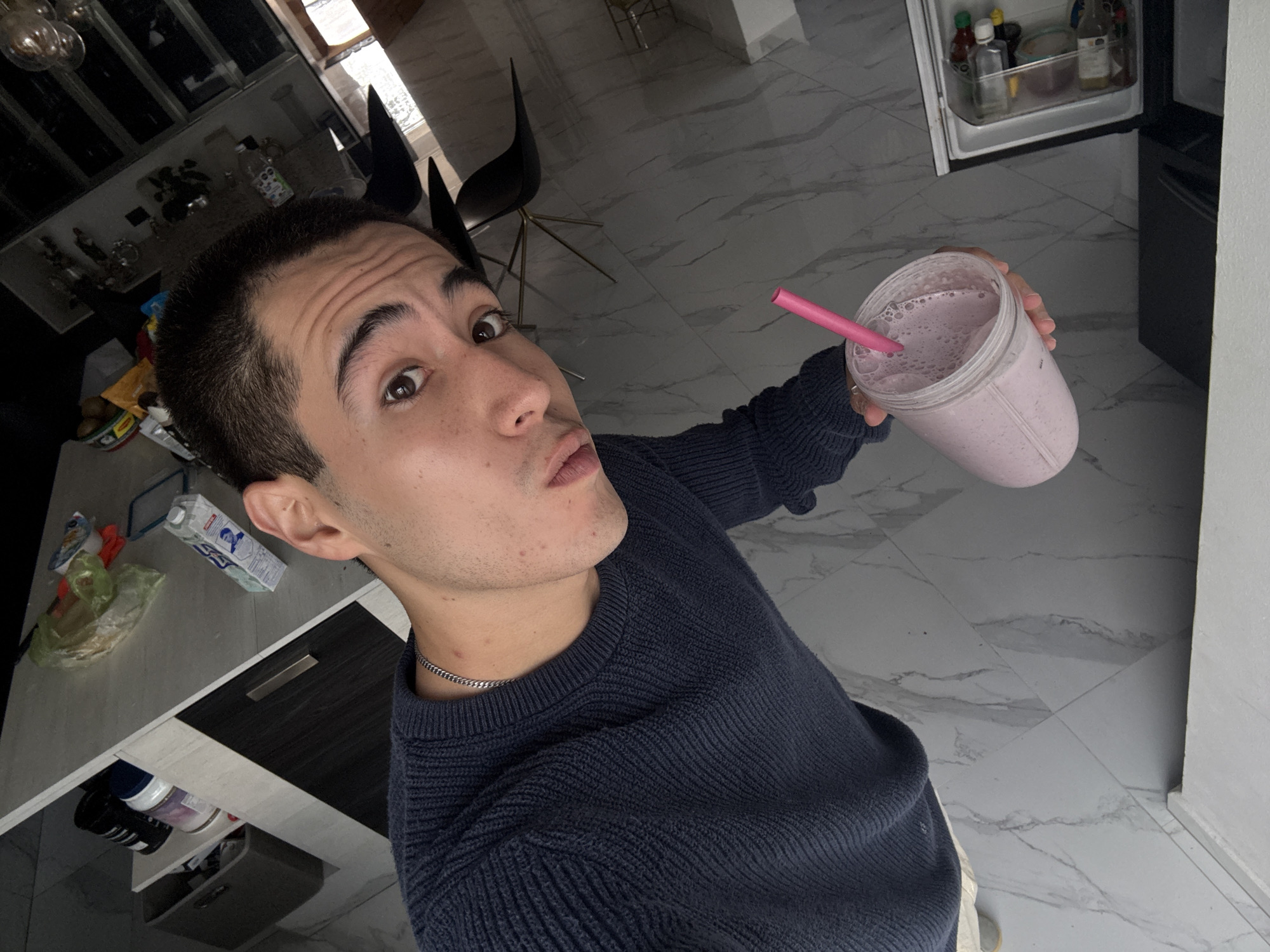 A young adult male is holding a smoothie with a pink straw in a residential kitchen. He has short dark hair and is wearing a navy blue sweater with a chain necklace. The kitchen features marble-like tile flooring, a black and white island with various items on top, and a refrigerator. A set of black chairs can be seen in the background. This image was taken in San Bernardino Tlaxcalancingo, Mexico.