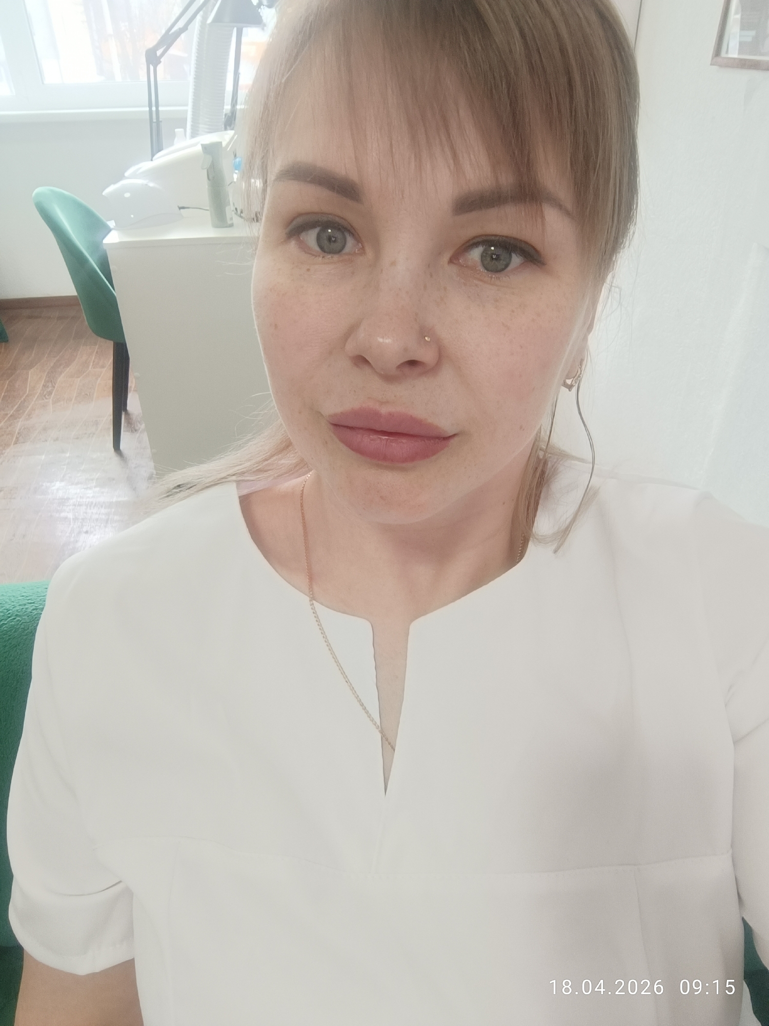 A woman is captured in a selfie-style portrait, looking directly into the camera with a neutral expression. She has fair skin with visible freckles, light-colored hair with bangs, and light green or gray eyes. A small, shiny piercing is visible on her nose. She is wearing a white, V-neck professional-style top or tunic, along with a delicate gold chain necklace. A distinctive long, wavy earring dangles from her right ear.
The setting appears to be an indoor professional environment, possibly a beauty salon or clinic, located in Chaikovsky, Russia. The background is brightly lit, with a window visible on the upper left. Behind her, to the left, is a white cabinet or desk supporting various pieces of equipment, including an adjustable lamp and other white devices that suggest a workspace for beauty services. A teal or dark green chair is partially visible behind the furniture. The floor features a light-toned patterned surface, possibly wood or laminate.
A digital timestamp "18.04.2026 09:15" is visible in the bottom right corner, indicating the date and time the photo was taken in the morning. The overall impression is one of a clean, well-lit professional space.