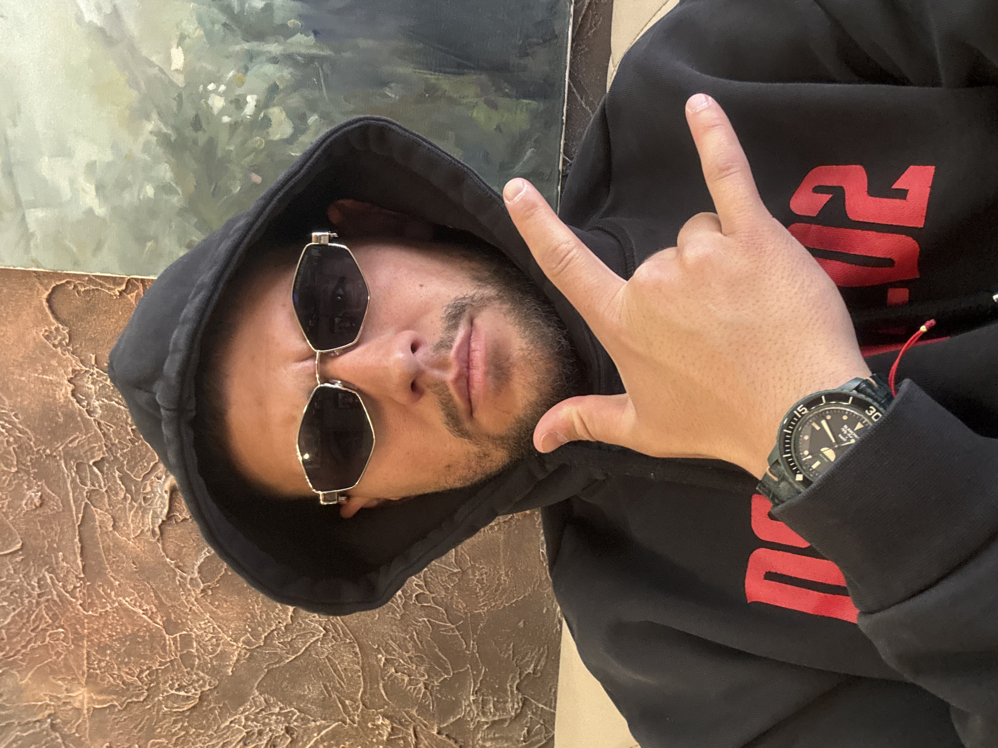 A person with medium skin tone, wearing a black hoodie with red lettering, is posing for a photo. The person has a short beard, sunglasses and a watch on their left wrist. The person is making a hand gesture. The background appears to be a wall with textured paint or plaster, and what might be a painting or decorative panel. It is not possible to determine the location in Sofia, Bulgaria, from the image alone.