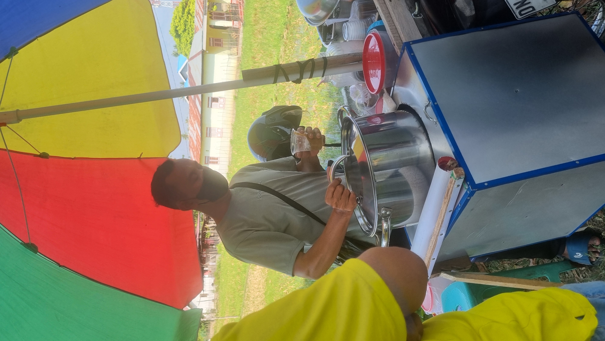An unidentified man wearing a grey t-shirt and a black face mask is standing under a large, colorful umbrella with red, yellow, and green sections. He is holding a clear plastic cup with a brown liquid in his right hand and is in the process of pouring something into a large metal pot with a silver lid. He has a black strap across his chest. To his left, a person wearing a bright yellow shirt is partially visible. The scene appears to be outdoors, possibly on the side of a road, with a grassy field and several small buildings in the background. A black motorcycle helmet rests on the edge of a cart or stand that holds the pot and other containers. The cart is made of metal with blue trim. A license plate that reads "ON" is visible on the right side of the cart. The overall impression is of a street vendor preparing a drink or food item.