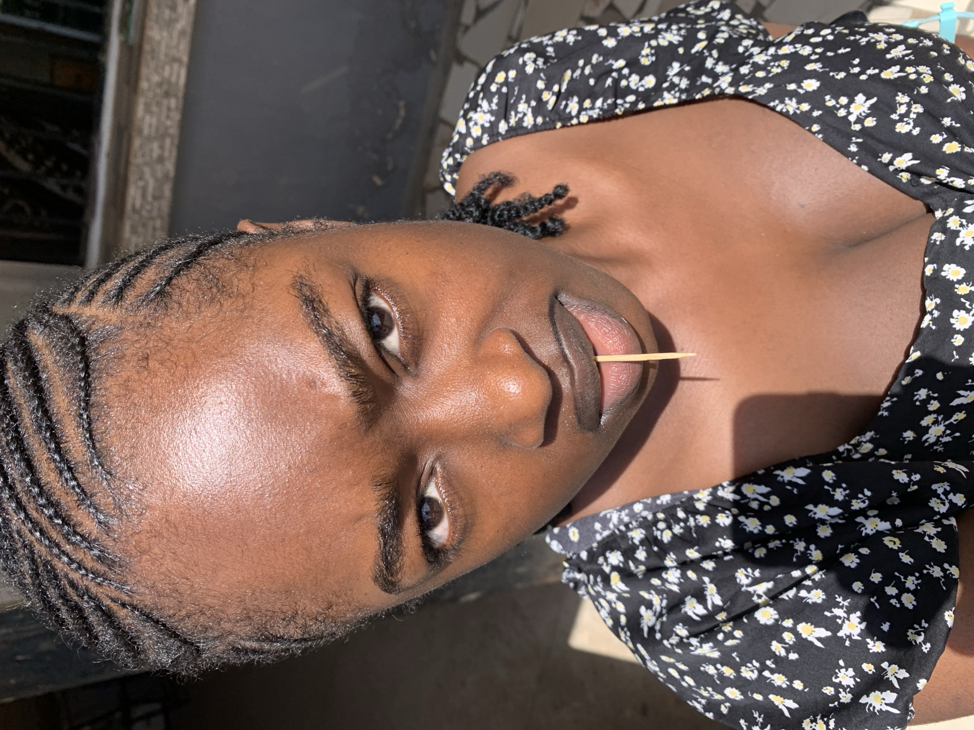 The image presents a diagonally oriented, close-up portrait of a young person with a dark skin tone, captured outdoors in bright sunlight at New Stadium, Nigeria. The subject is looking directly towards the camera with a neutral expression. Their hair is styled in neat cornrows on the scalp, with additional short, coily braids visible around the neck. A slender wooden toothpick is held between their lips.
The person is wearing a black top with a repeating pattern of small white and yellow flowers, which is visible across their upper chest and shoulders. Strong sunlight illuminates their face and skin, creating highlights and suggesting a clear, sunny daytime environment. The background is out of focus, showing indistinct architectural elements, possibly parts of a building or wall structures, typical of an outdoor setting. No text is visible within the image.
