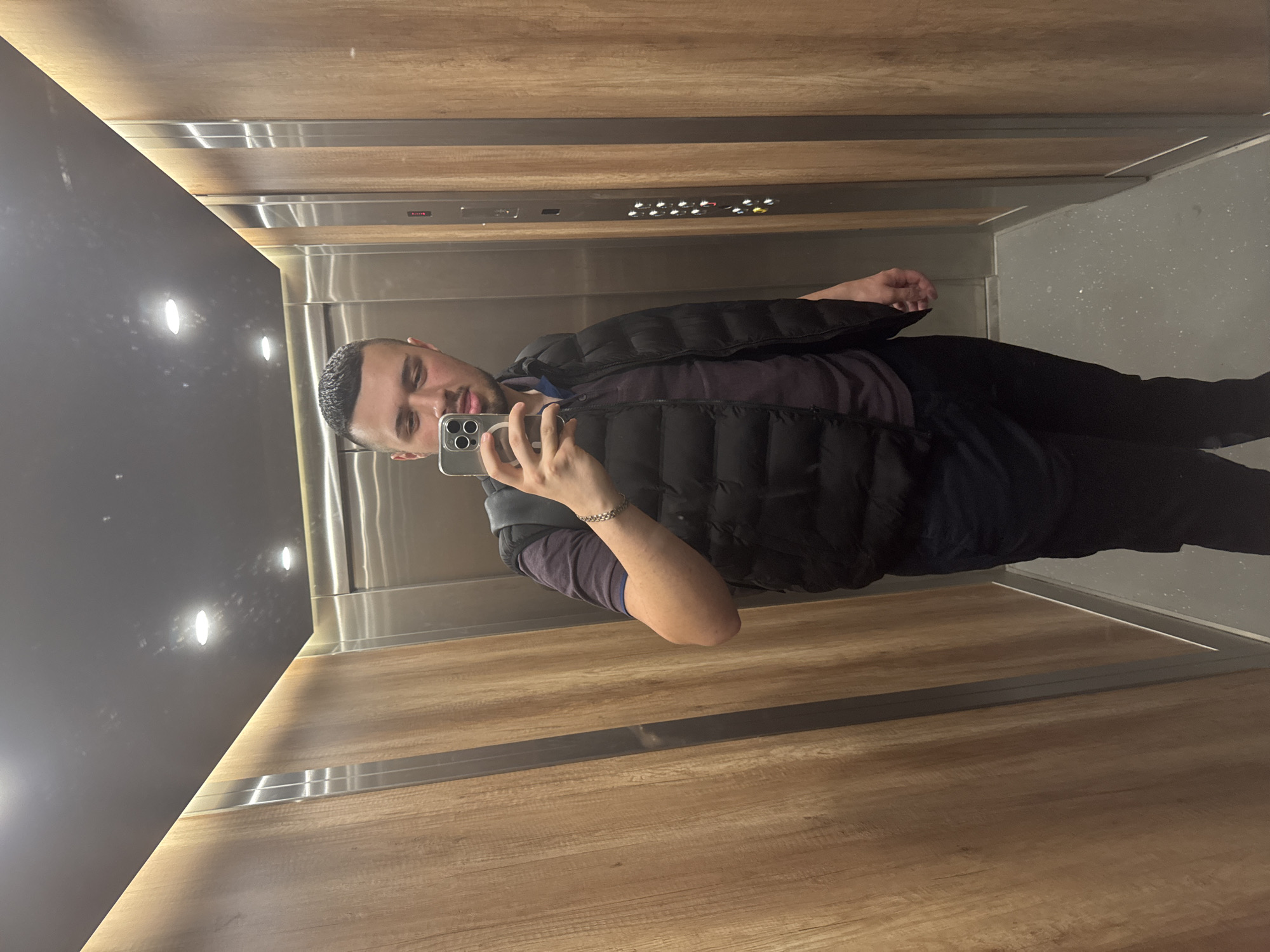 A man takes a selfie in an elevator. He is wearing a black puffer vest over a dark shirt and black pants. He is holding a silver iPhone. The elevator has wood paneling on the walls, a dark ceiling with recessed lighting, a stainless steel door, and gray flooring. There are elevator control buttons on the wall. The image provides no information indicating that it was taken in Sofia, Bulgaria.