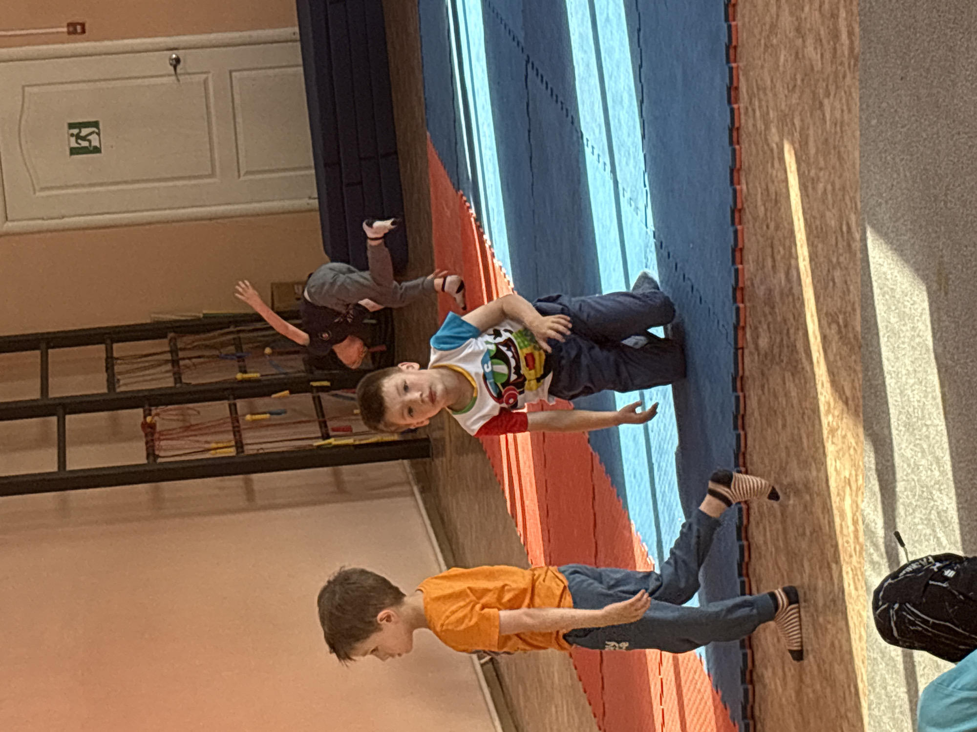 The image shows the interior of a sports hall, possibly a gymnastics facility, in Chaikovsky, Russia. In the foreground, two young boys are present. One boy, on the left, is wearing an orange t-shirt and blue pants, walking to the left. The other boy, in the center, is wearing a multi-colored t-shirt with cartoon characters and blue pants, kneeling on a blue mat. In the background, another child is in mid-air, performing a somersault or flip. The hall has a wooden floor covered with mats in red and blue sections. A large rack with various ropes and jump ropes is visible on the left. A white door with an emergency exit sign is on the right wall, and a fire alarm is mounted next to it. The lighting suggests it is daytime, with sunlight casting shadows across the floor.
