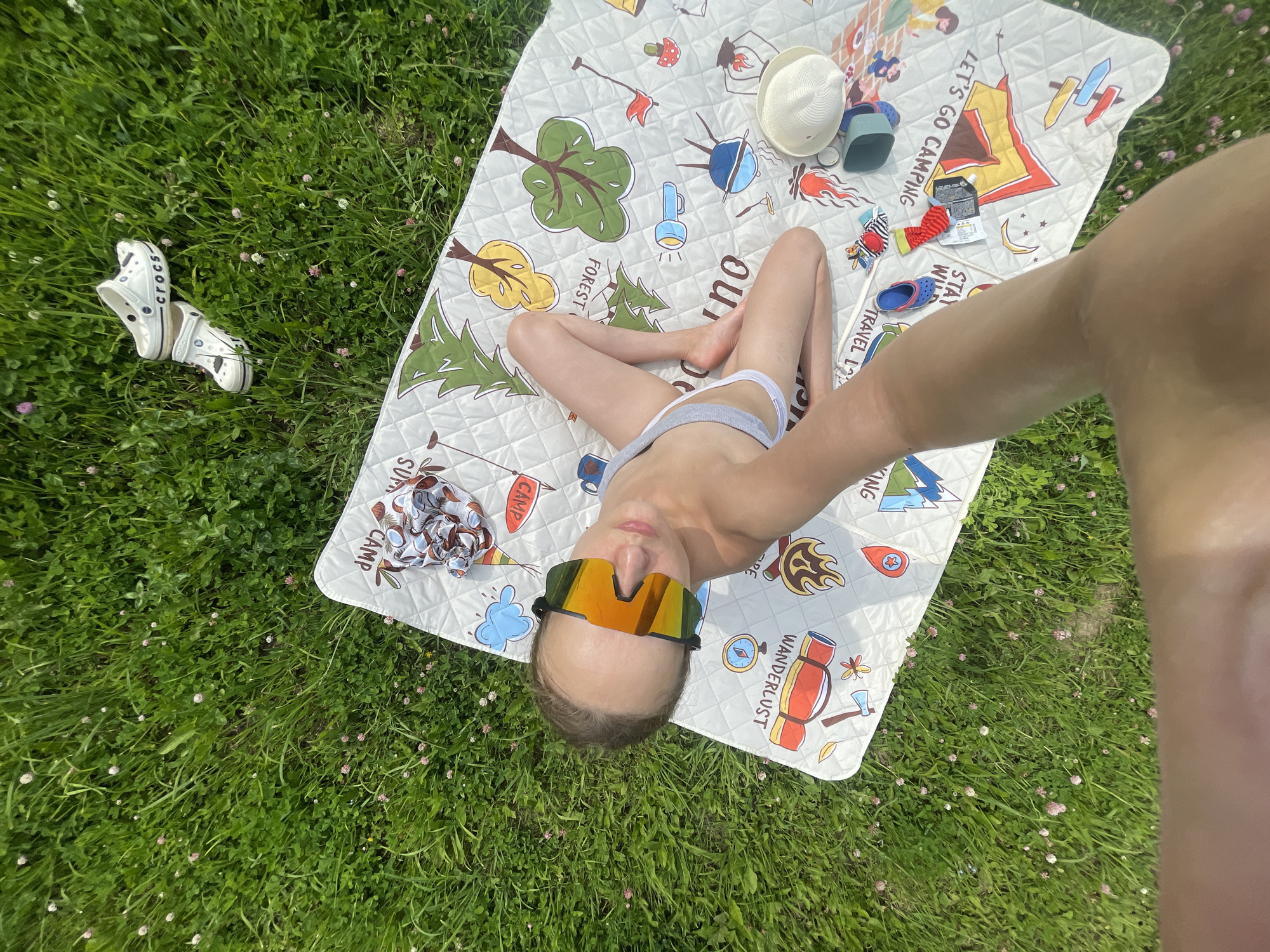A person lies on their back on a picnic blanket spread on a grassy lawn. The person is wearing sunglasses with orange lenses and grey underwear. The picnic blanket has a design featuring camping-related illustrations, including tents, trees, and outdoor equipment. Scattered around the blanket are a pair of white Crocs shoes, a white sun hat, a small blue container, and a pair of striped socks. The scene appears to be outdoors in a natural setting, likely a park or backyard, under daylight conditions. The grass is green and dotted with small pink and white flowers. Text visible on the blanket includes "LET'S GO CAMPING," "WANDERLUST," "STAY WILD," and "CAMPING." The overall mood is relaxed and leisurely, suggesting a peaceful outdoor activity.