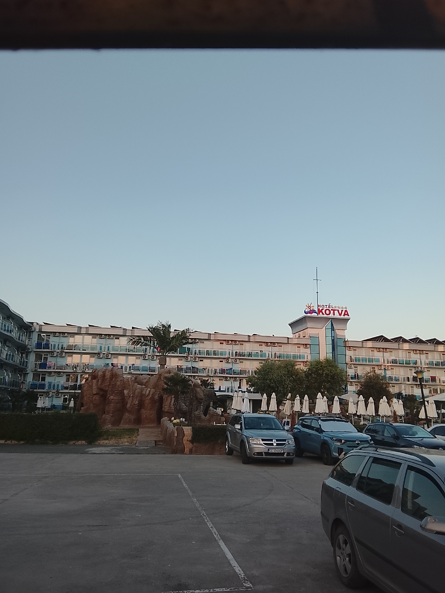 The image shows a parking lot in front of a hotel in Nesebar, Bulgaria. The hotel, named "KOTVA", has a distinctive red sign with a sun and waves graphic. The hotel building is multi-story with many balconies and air conditioning units. In the foreground, several cars are parked, including a silver SUV, a blue SUV, and a dark-colored sedan. To the left of the hotel, a man-made rock formation with a palm tree is visible. White umbrellas are arranged in rows behind the rock formation. The sky is clear and light blue, suggesting it is daytime.