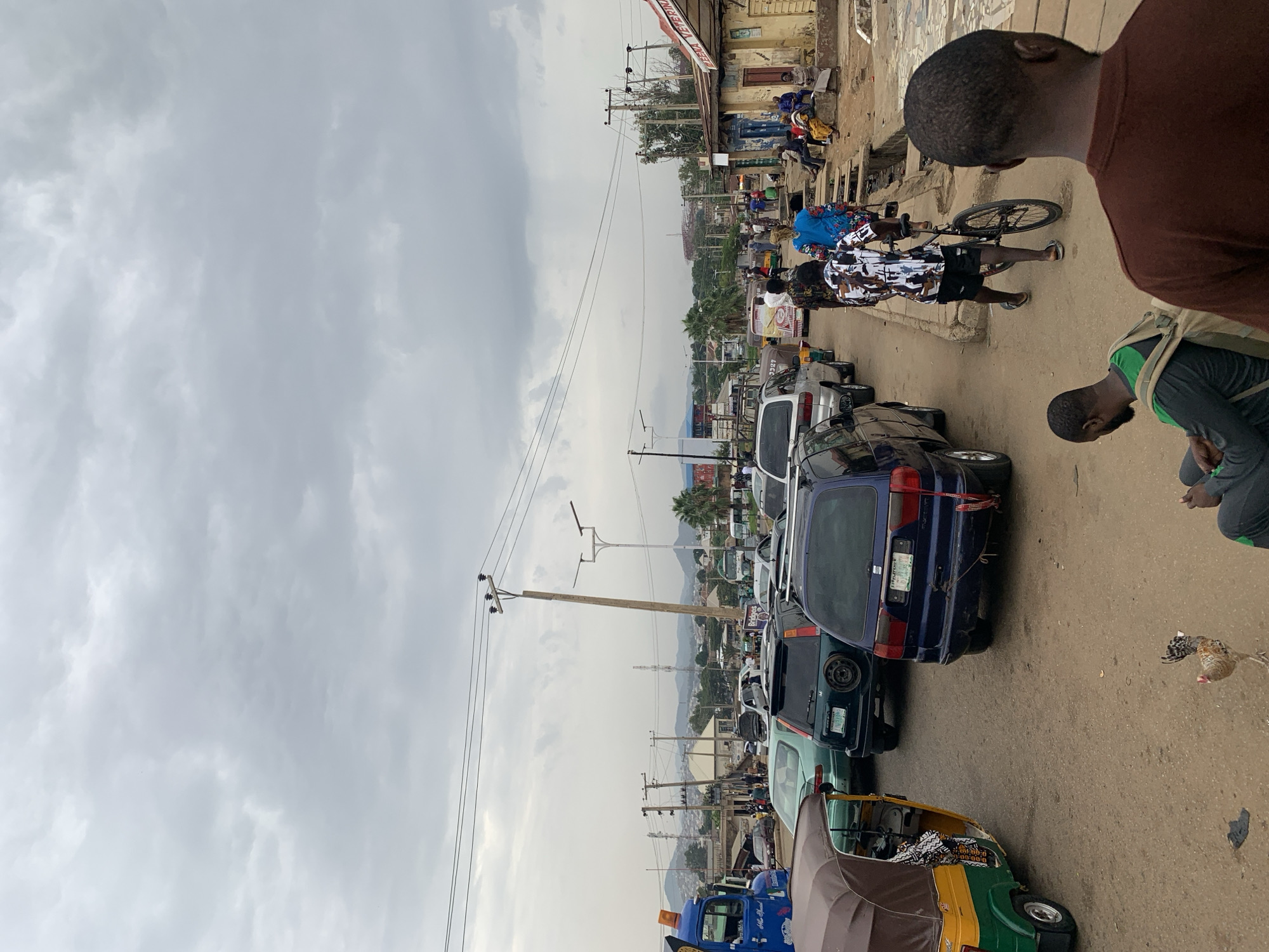 The image depicts a street scene in Rafin Par, Nigeria, characterized by a mix of vehicular and pedestrian traffic. The foreground features a row of parked or slow-moving vehicles, including a blue car with visible license plates, a green car, and a three-wheeled auto-rickshaw.
Several individuals are visible. One person is riding a bicycle, while others are walking alongside the vehicles. One person is seen bent over, and another is visible from the back, partially cut off by the image frame. The environment includes utility poles with power lines extending overhead, and buildings are visible in the background, suggesting a bustling commercial area. The sky is overcast with clouds.