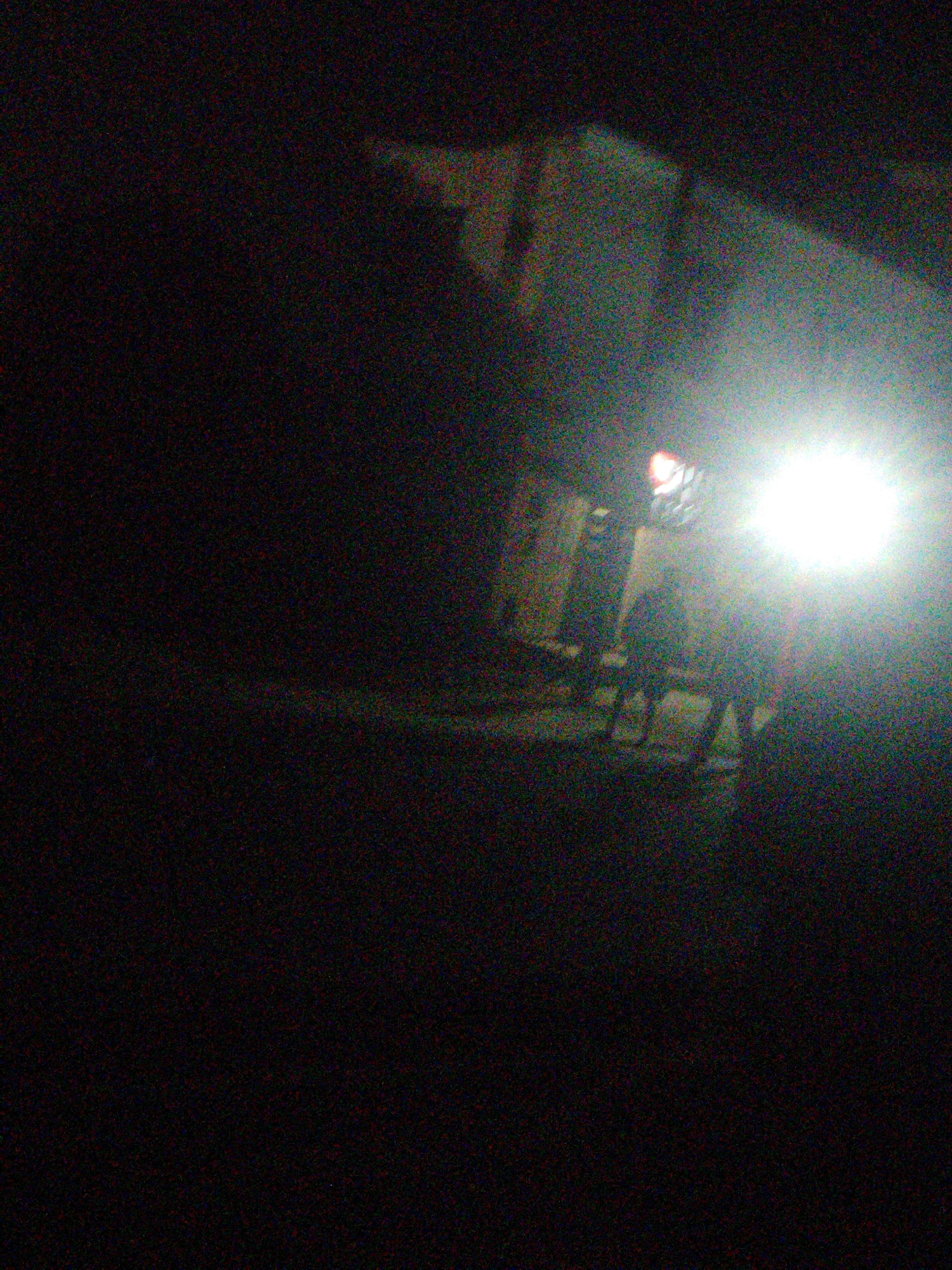 The image depicts a dimly lit outdoor scene, possibly at night, with a bright light source illuminating the right side of the frame. In the center of the image, there are several indistinct figures, some appearing to be standing, and others possibly sitting on chairs or benches. The figures are silhouetted against the light and lack clear detail. In the background, there are faint outlines of structures, suggesting an urban or semi-urban environment. The image quality is poor, with significant noise and blurriness, making it difficult to discern specific features. The scene does not provide enough detail to definitively identify a specific location within Uyo, Nigeria, though the presence of structures and potential human activity is consistent with an urban setting.